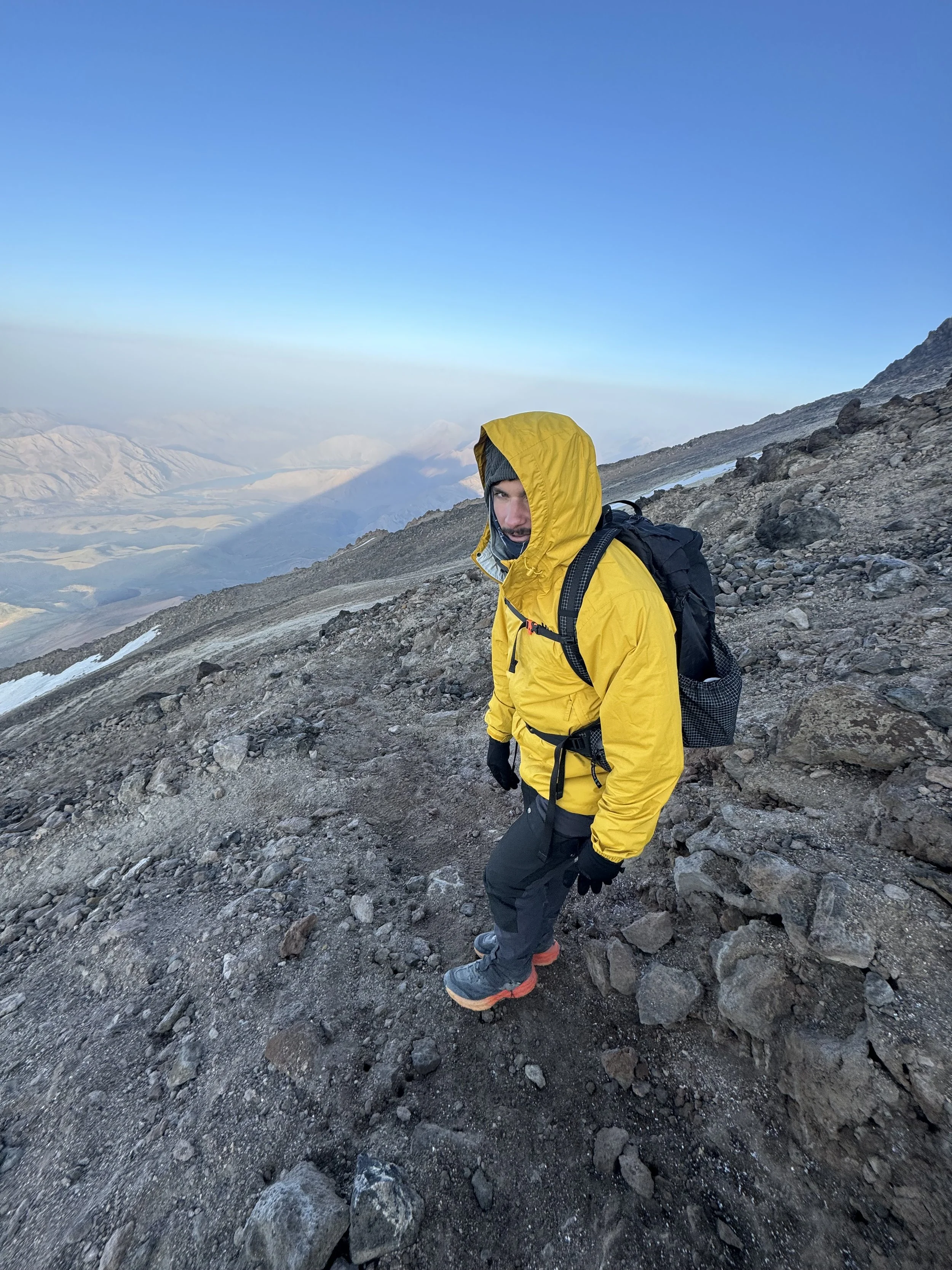 A person hiking on a rocky mountain trail wearing a yellow waterproof jacket, gray hood, black gloves, and black pants with orange soles. The person has a black backpack and is looking at the camera, with a mountain landscape in the background under