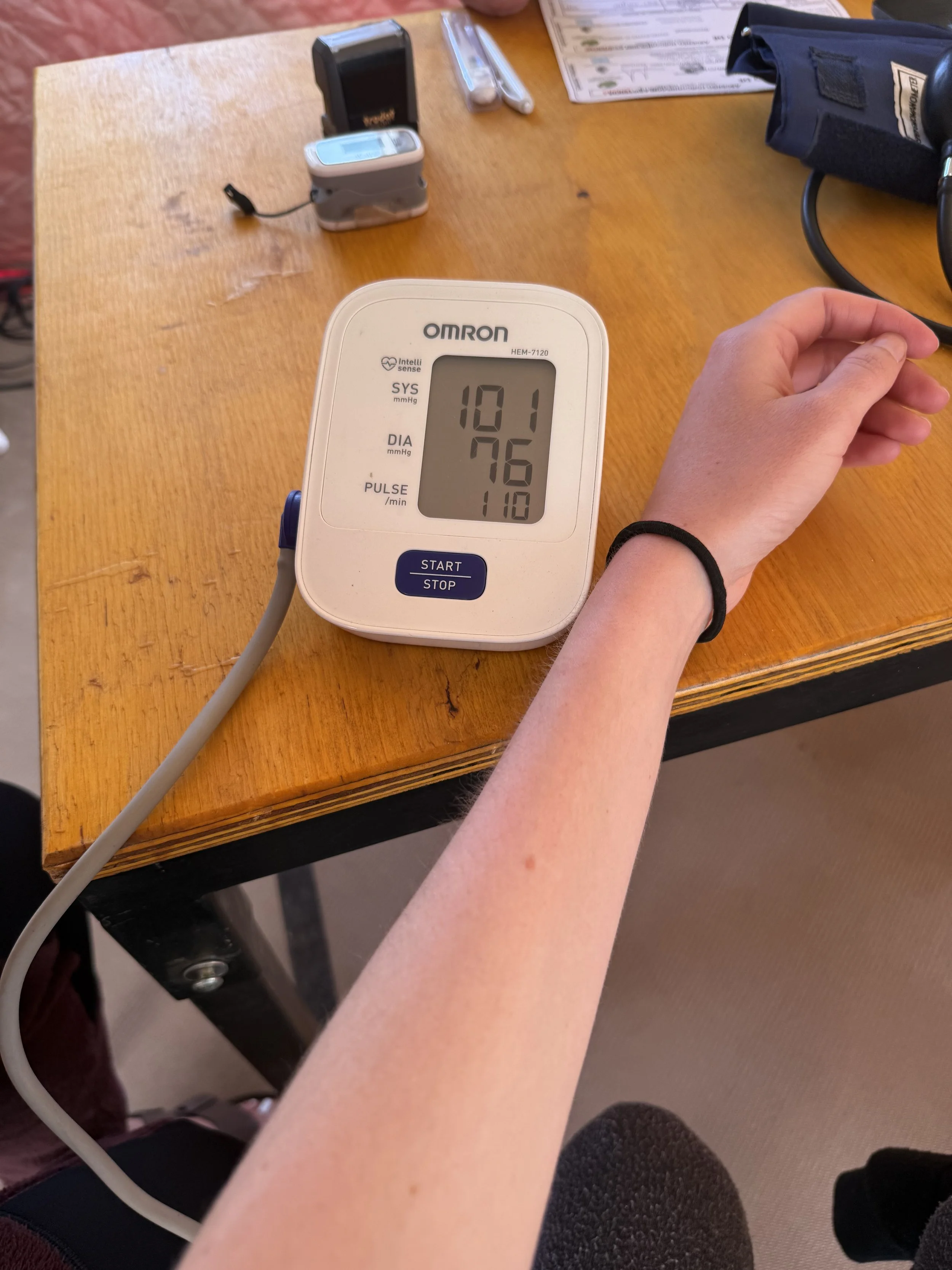 A person's arm resting on a table with an automated blood pressure monitor displaying a reading of 101 over 76 with a pulse rate of 110. The cuff is wrapped around the person's arm.