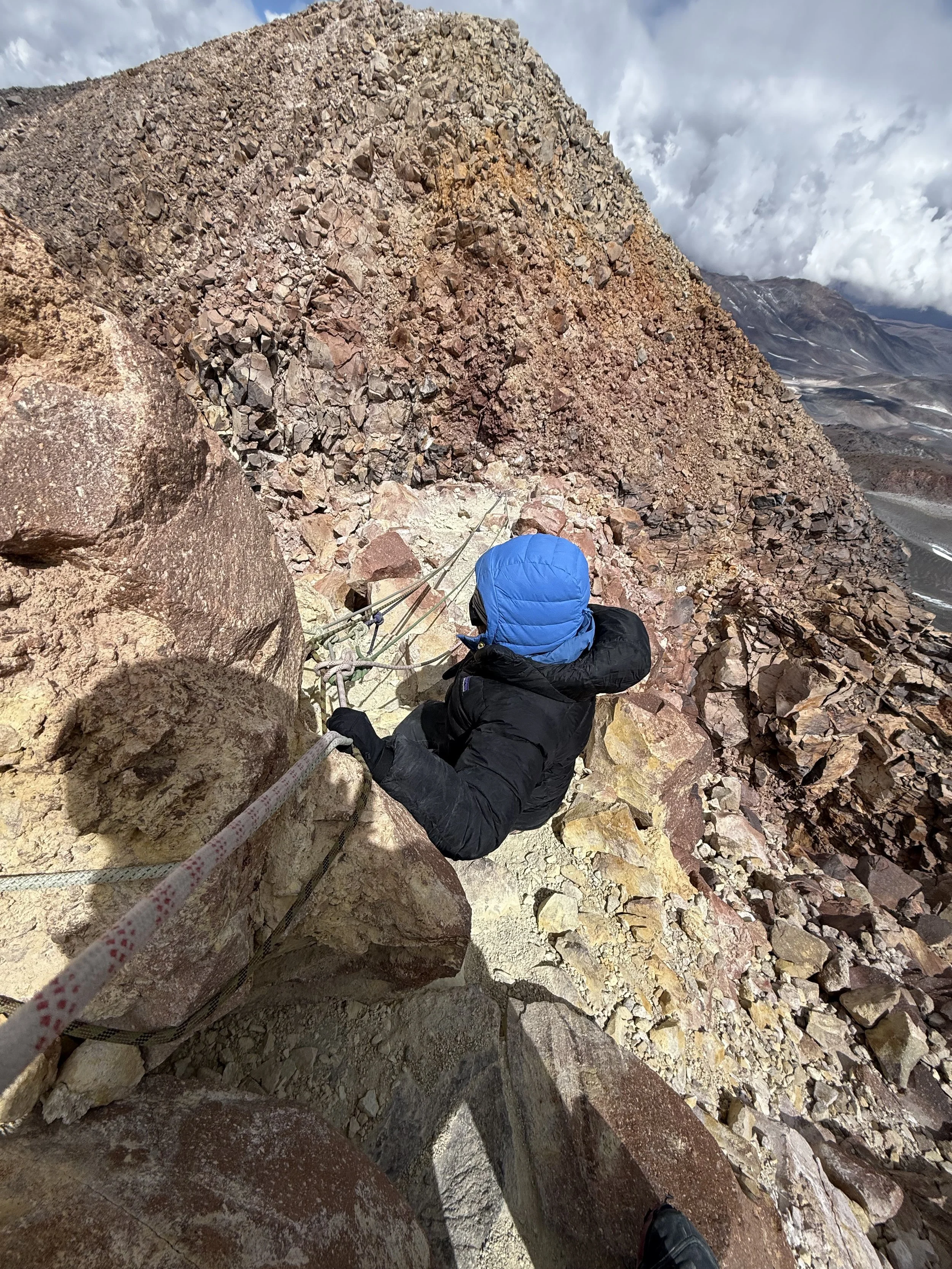A person climbing a steep rocky mountain, secured with ropes and wearing a blue hooded jacket and black gloves.