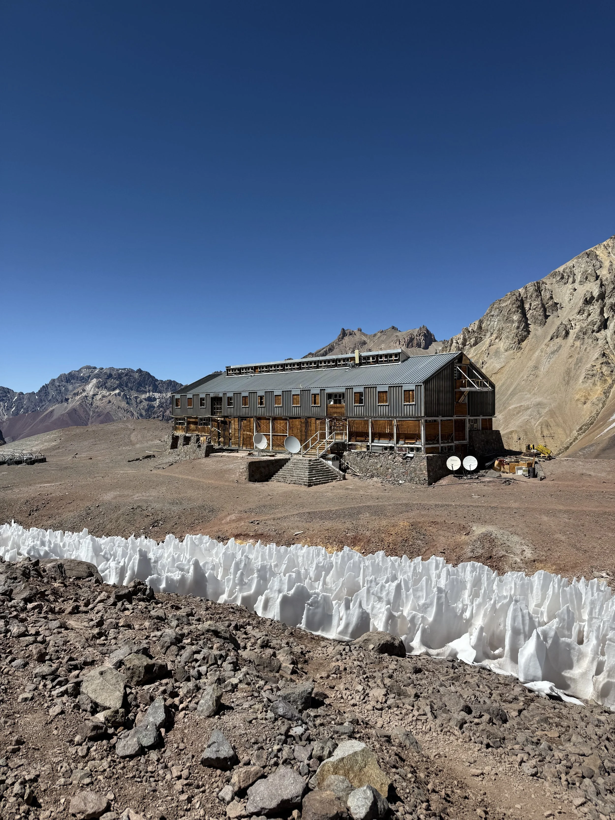A rugged, metal building situated on a barren, rocky landscape with brown and reddish mountains in the background under a clear blue sky.