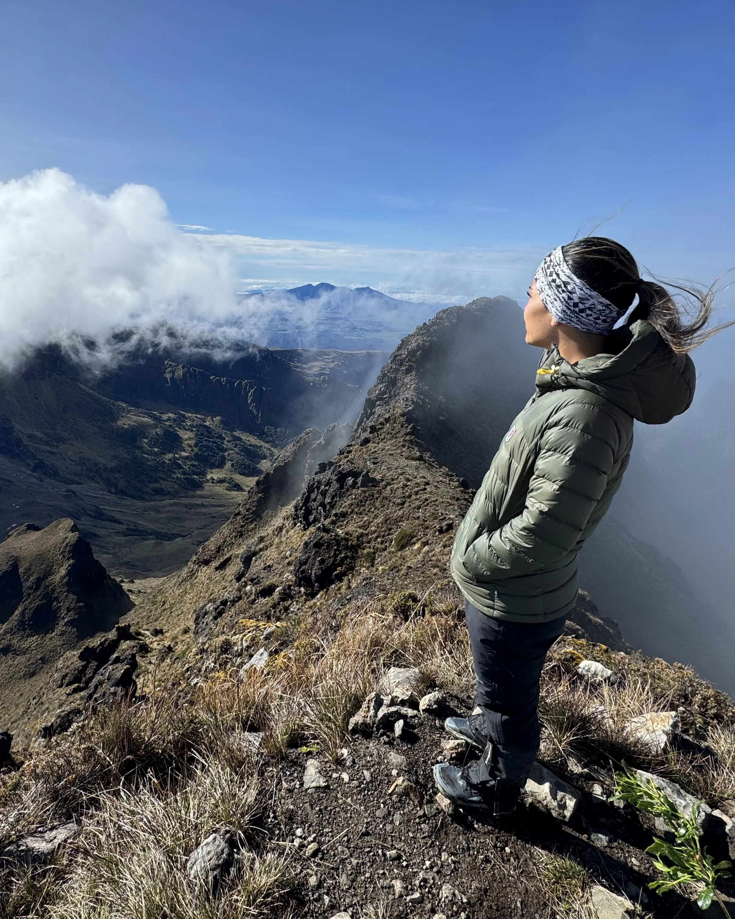 A woman dressed in outdoor hiking gear standing on a mountain ridge, looking at a landscape of mountains with clouds and blue sky in the background.
