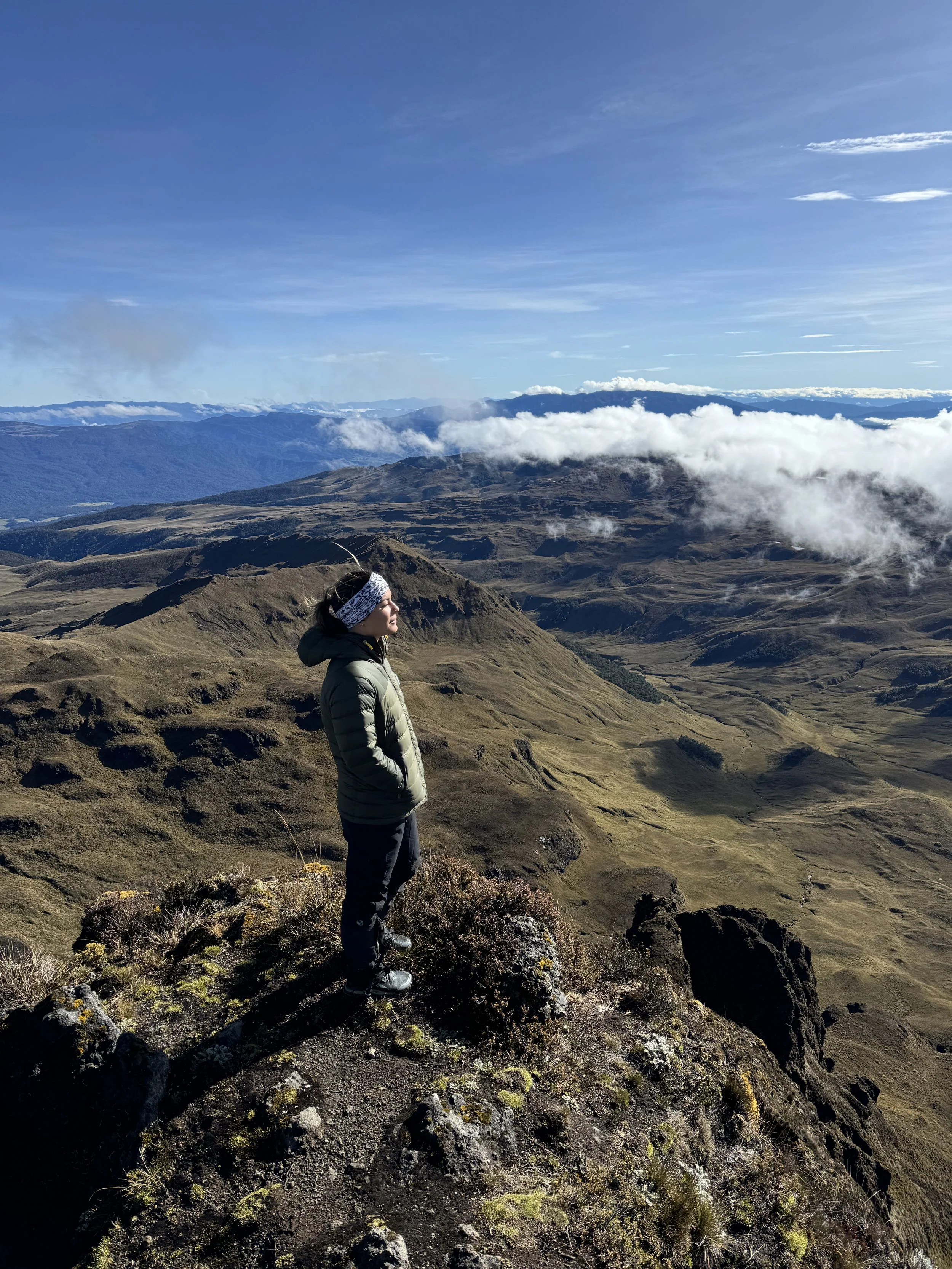 A woman standing on a rocky mountaintop with a view of rolling hills, mountains, and clouds in the distance under a blue sky.