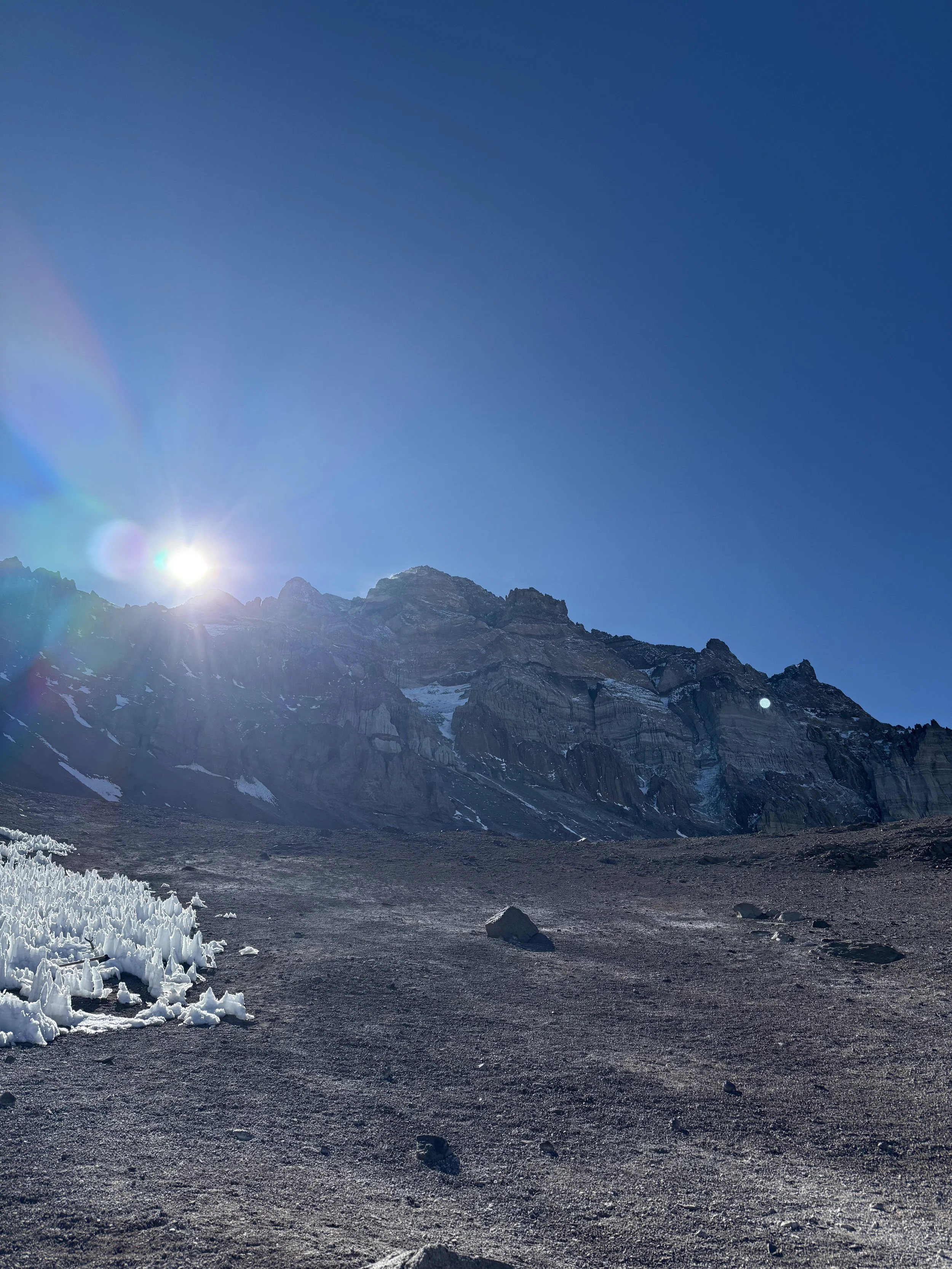 Scenic mountain landscape with a bright sun in a clear blue sky, snow and ice on the rocky terrain in the foreground.