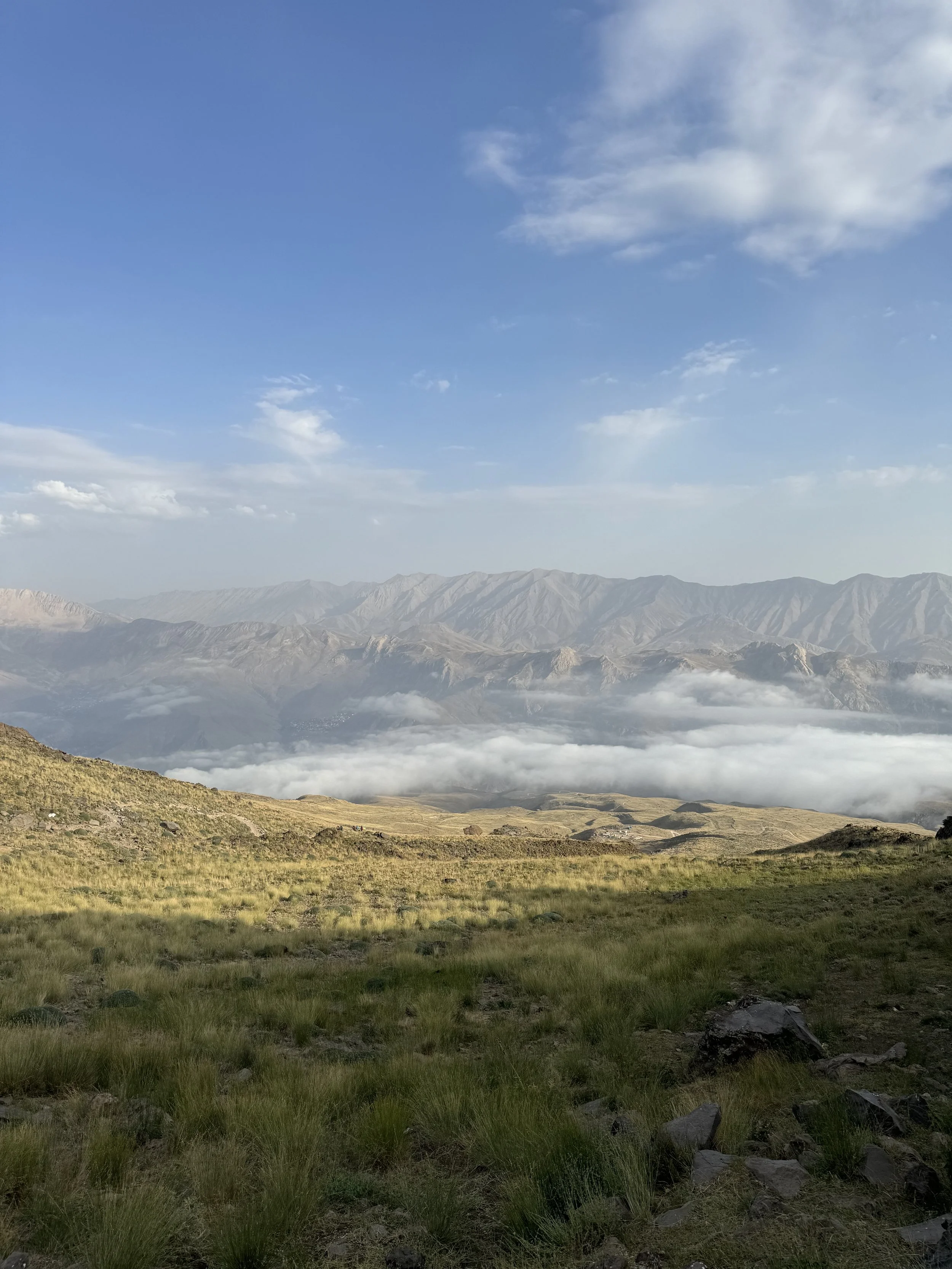 Mountain landscape with rolling hills, cloudy fog in the valley, and distant mountain range under a partly cloudy blue sky.
