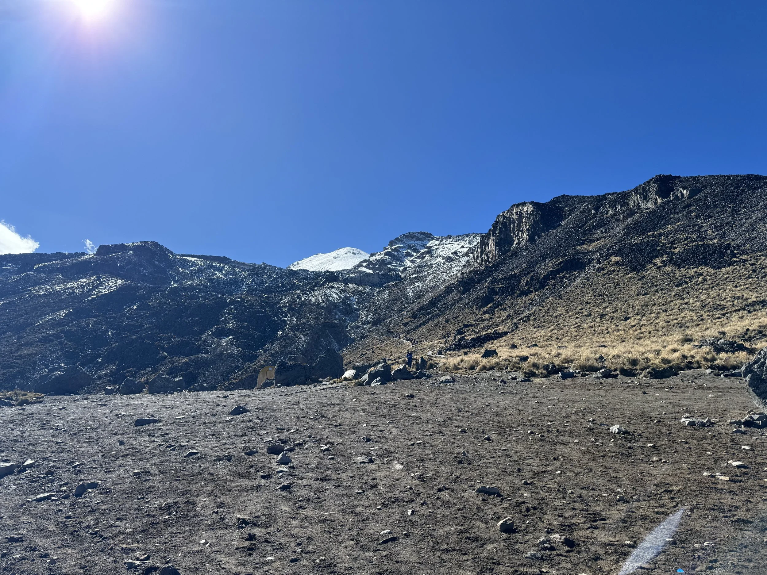 A mountainous landscape with a clear blue sky, snow-capped peaks, rocky slopes, and a dirt trail in the foreground.