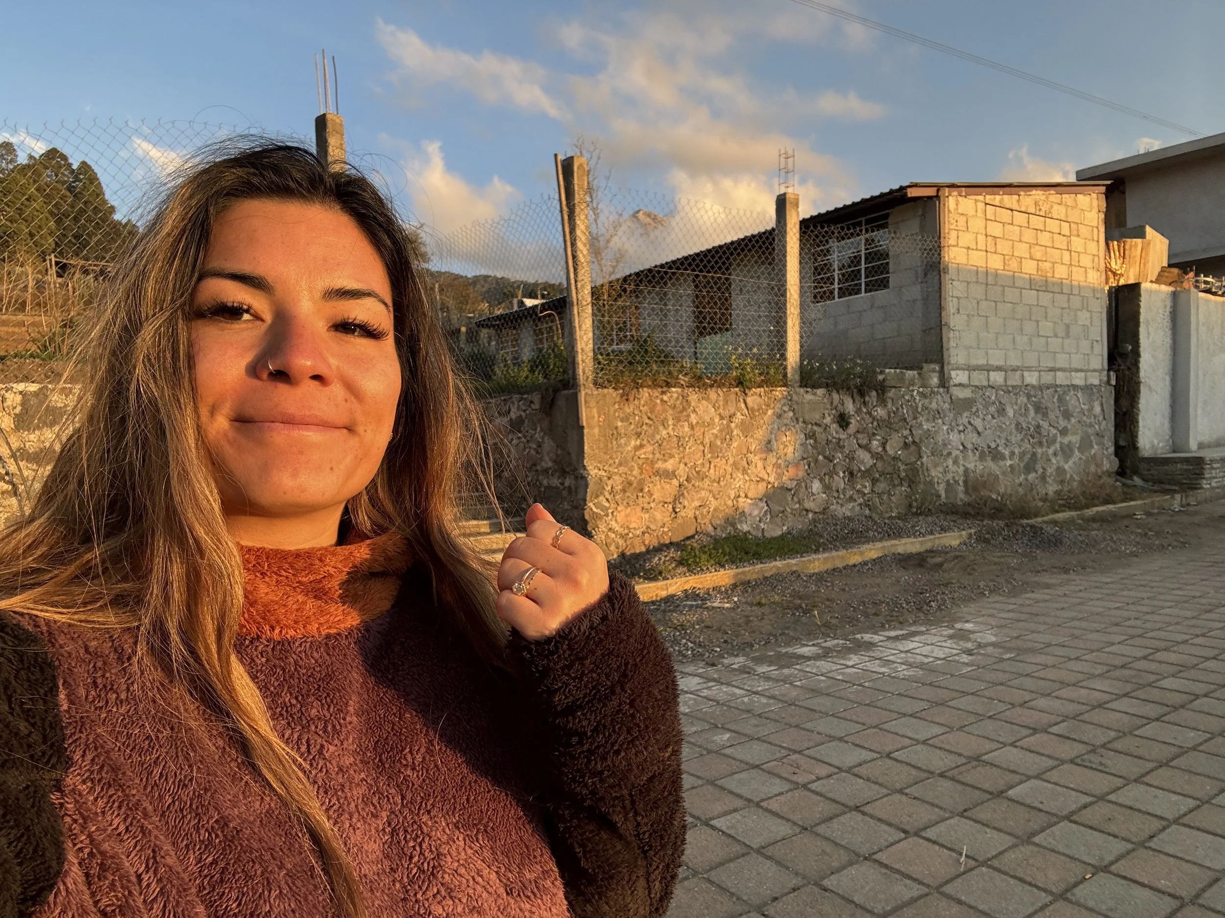 A woman with long hair, wearing rings and a nose piercing, taking a selfie during sunset, with a background of a stone wall, houses, and a partly cloudy sky.
