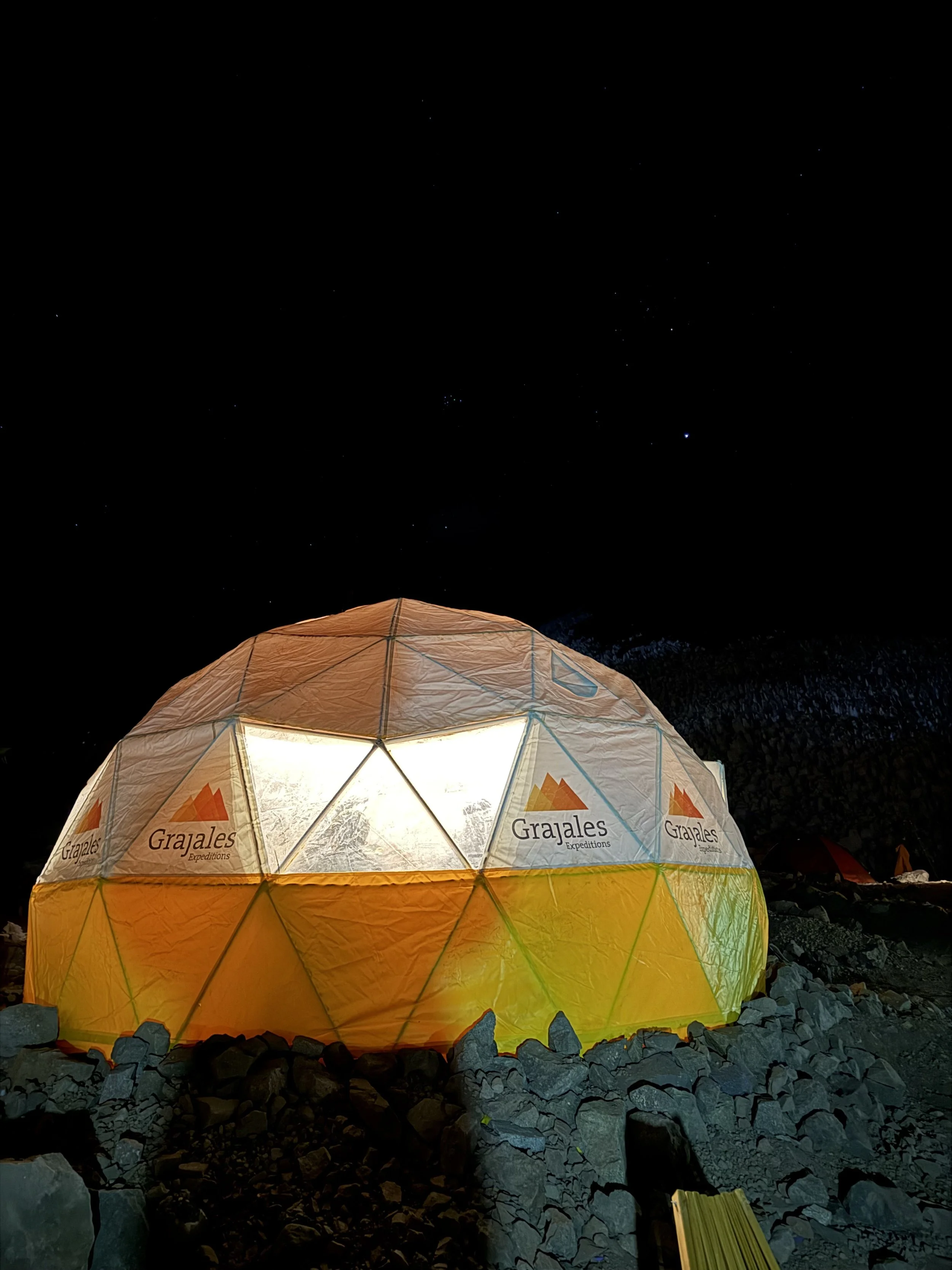 A geodesic dome tent illuminated from within, set against a dark night sky with stars. The tent has the logo and name "Grajales Expeditions" printed on it, and is surrounded by rocks and smaller tents.