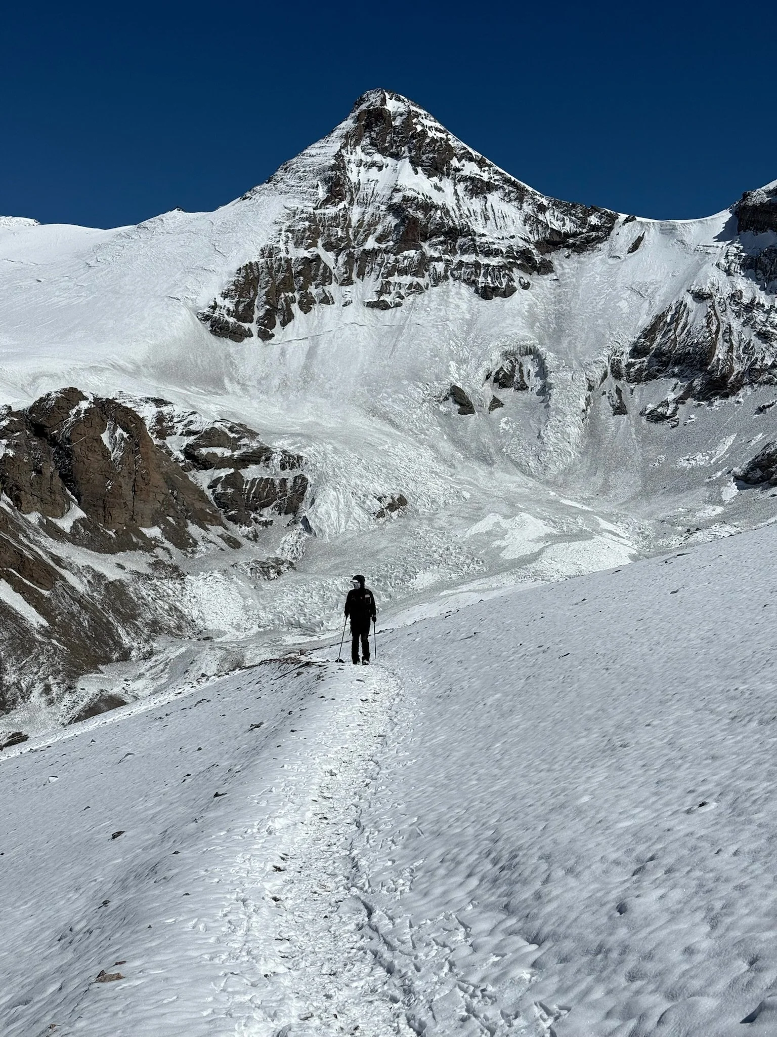 A person hiking on a snow-covered trail in a mountainous, glacial landscape with a tall, snow-capped peak in the background.