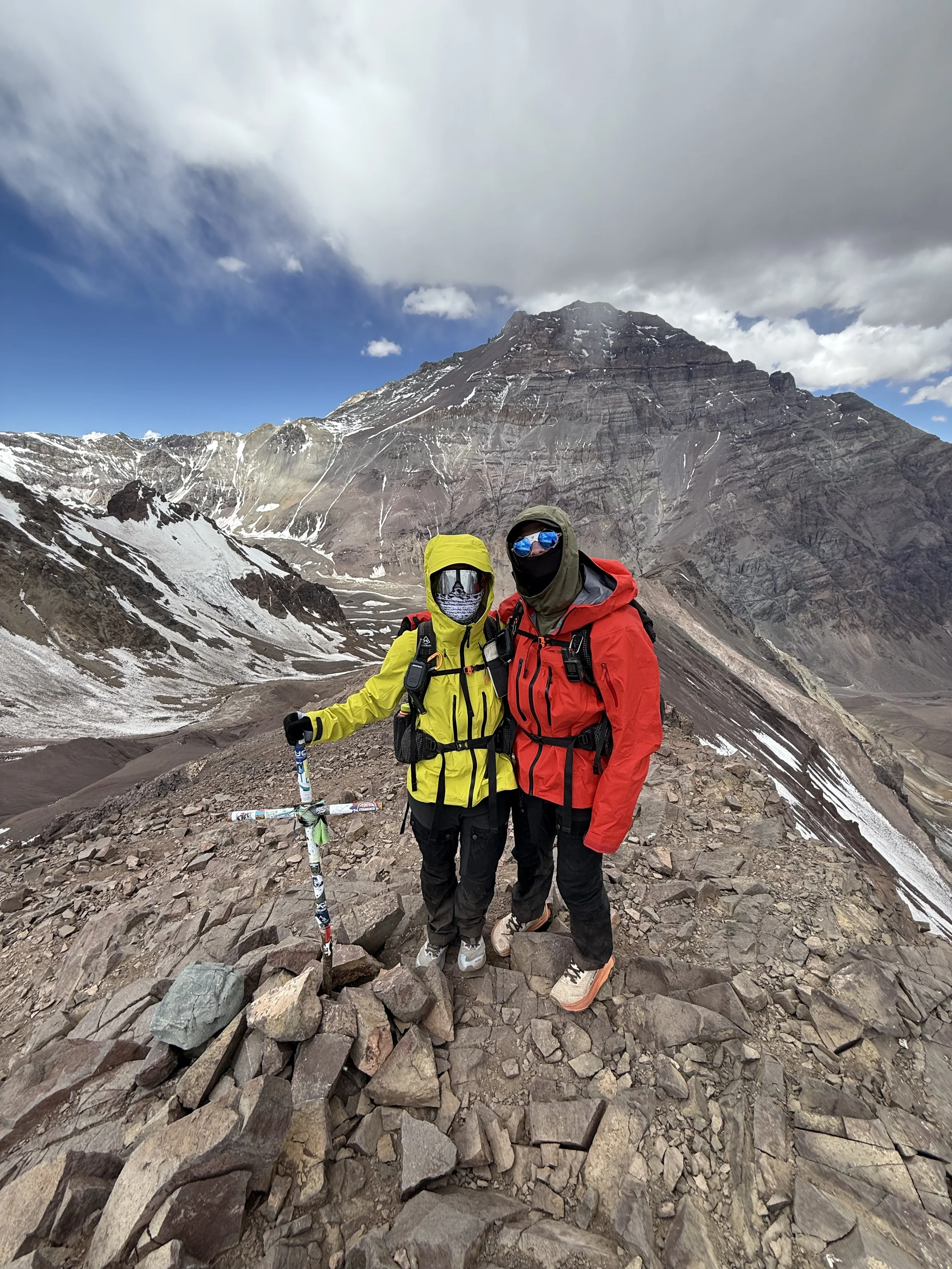 Two mountaineers in bright jackets and protective gear standing on rocky terrain in a mountain landscape with snow patches and a towering mountain in the background.