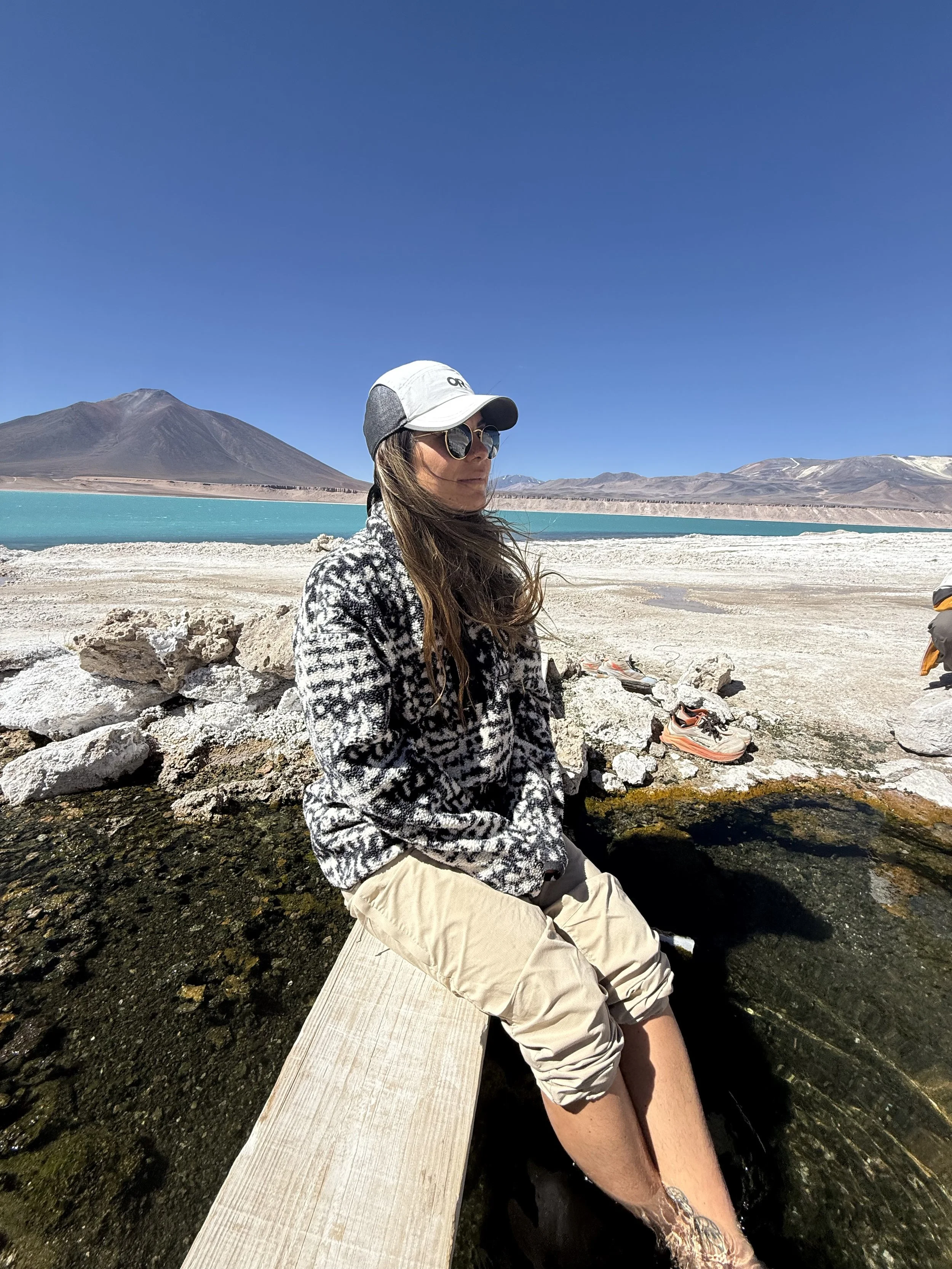 A woman sitting on a wooden bench with her feet in a hot spring, with a mountain, lake, and clear blue sky in the background.