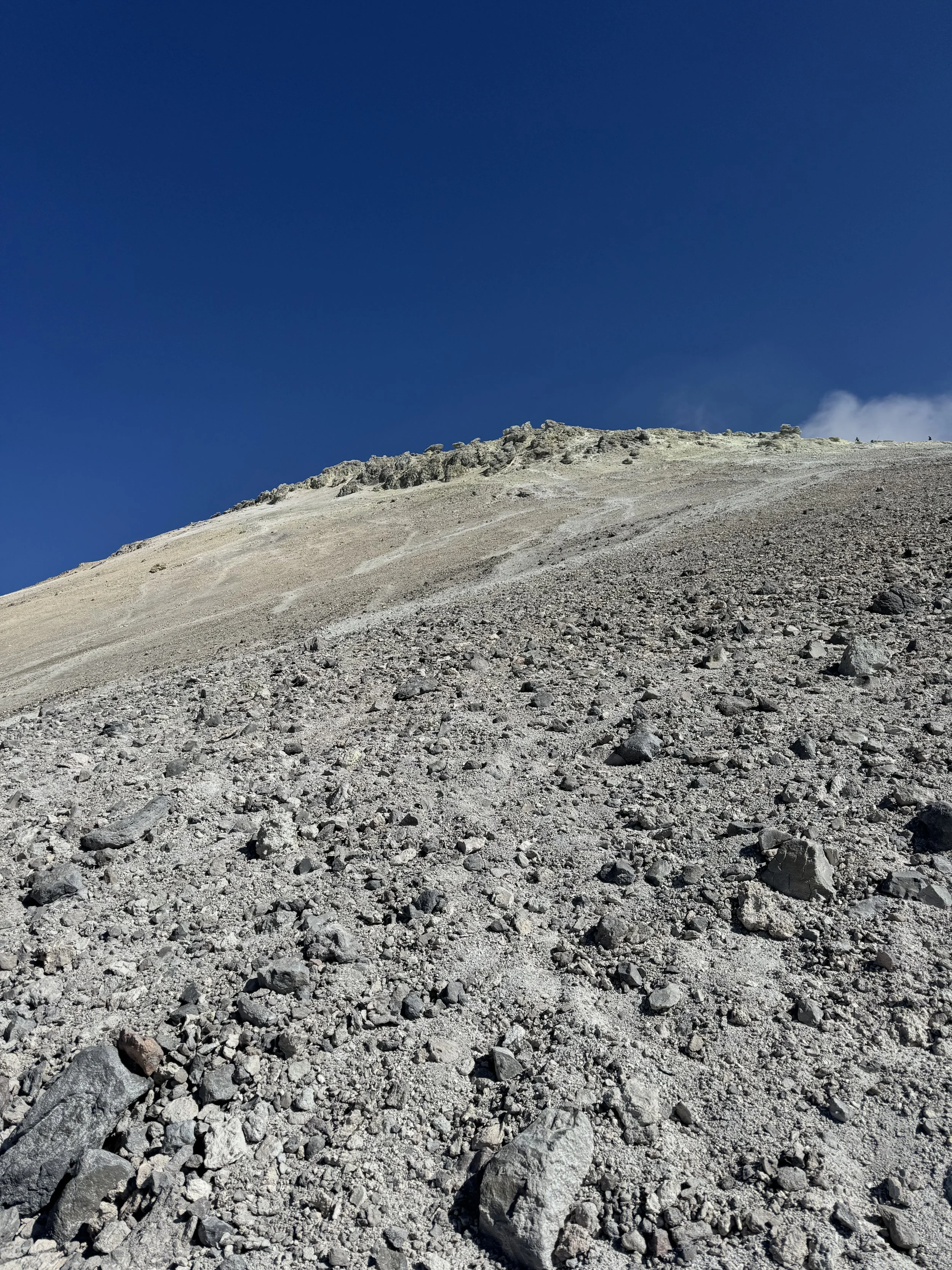 A rocky mountain slope with loose gravel and stones leading up to a ridgeline under a clear blue sky.
