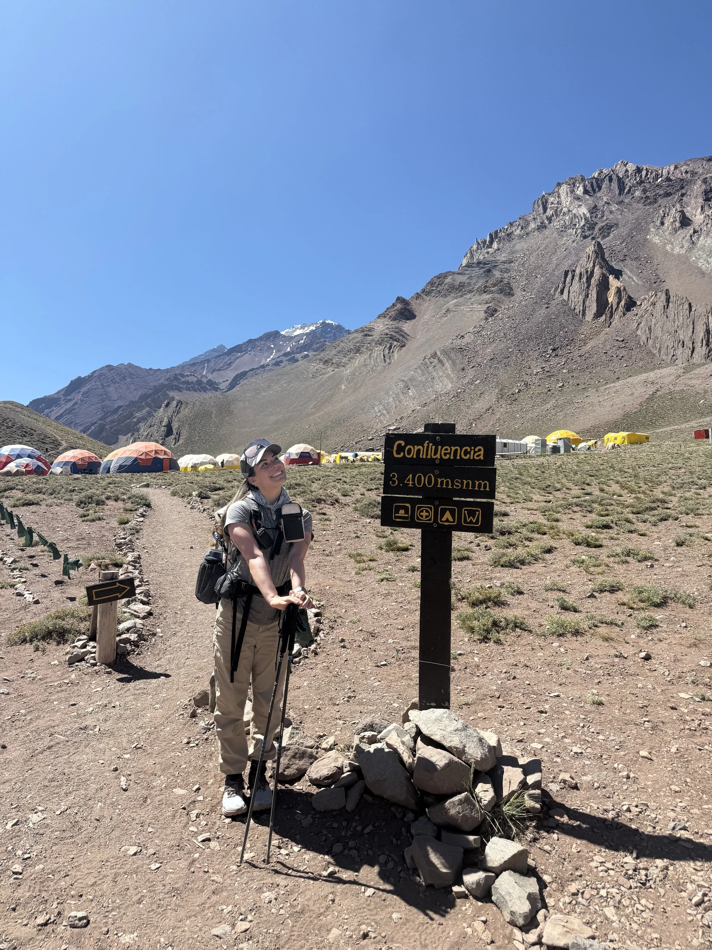 A woman with hiking gear standing next to a sign at Confluencia, 3400 meters above sea level, with tents and mountain landscape in the background.