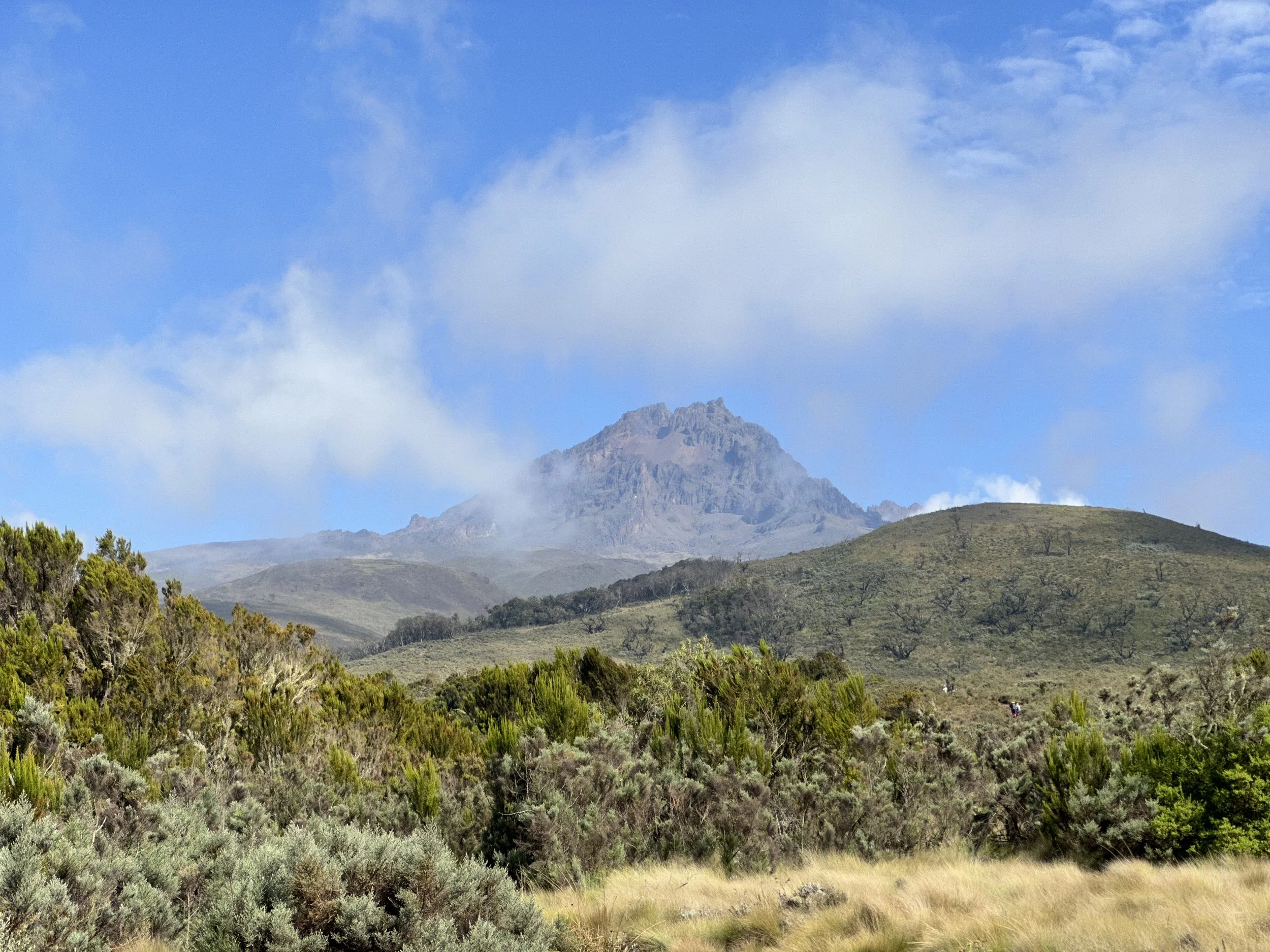 Landscape view of a mountain with some clouds in the sky, surrounded by green shrubs and rolling hills.