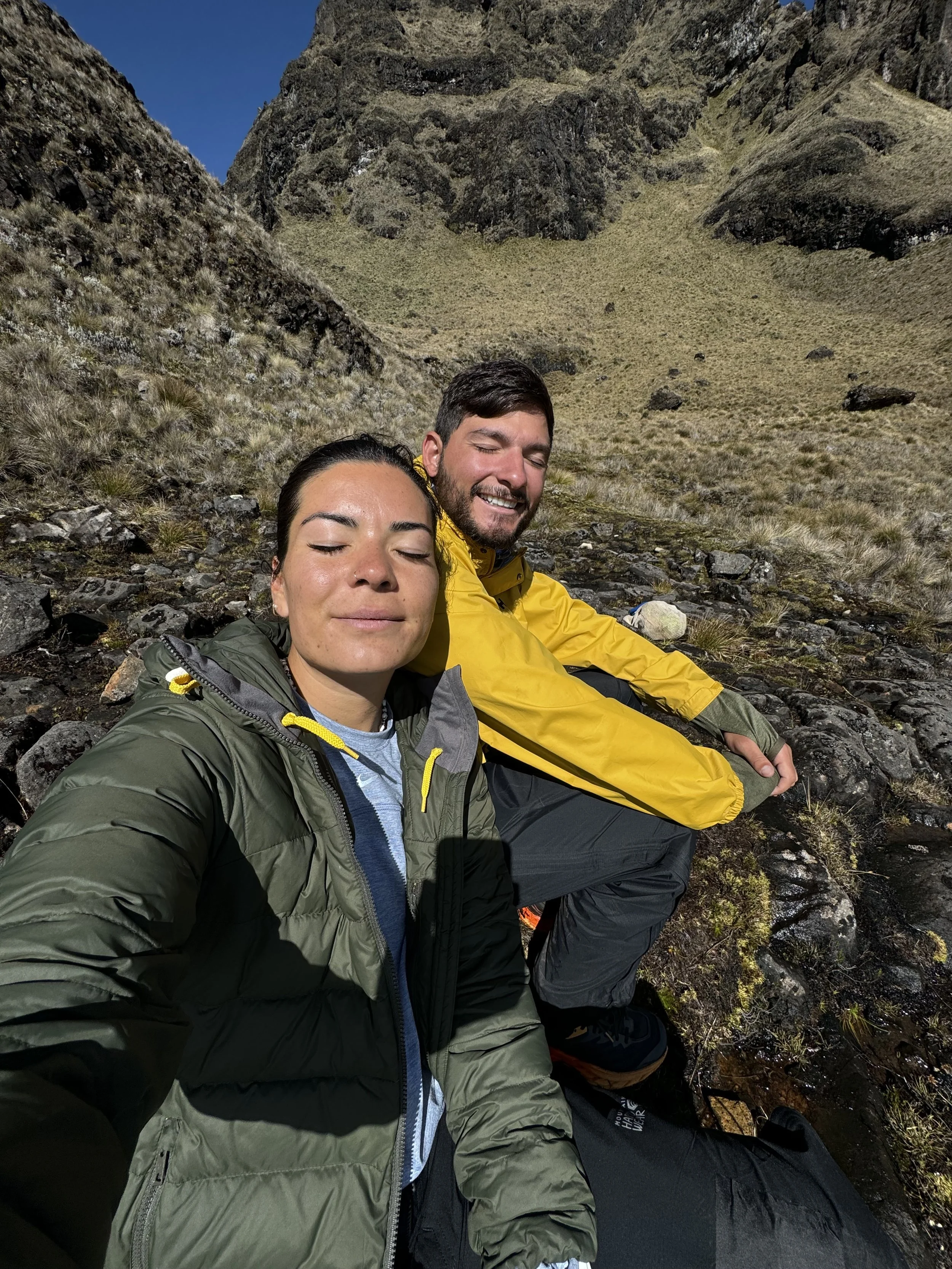 A woman and man sitting on rocks in a mountainous outdoor setting, both wearing jackets, smiling with closed eyes, enjoying the sunlight.