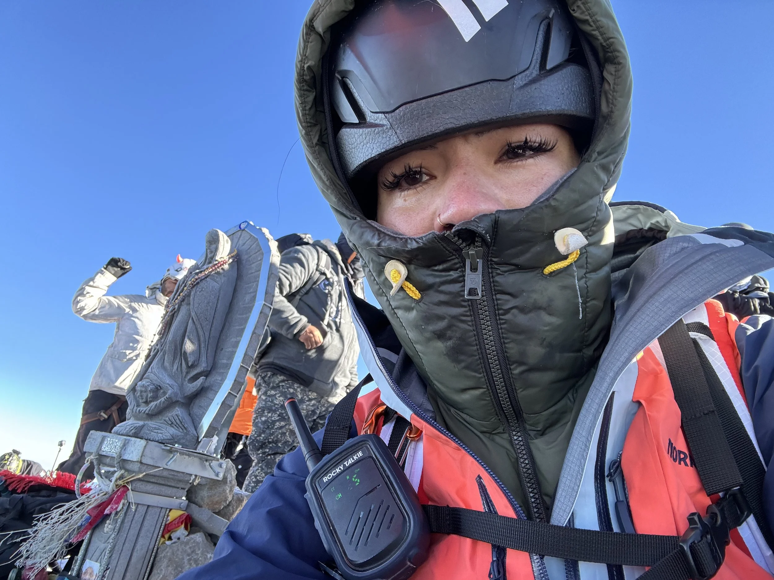 Close-up of a woman in mountain gear, wearing a helmet and high-altitude clothing, with others in similar gear behind her, all on a mountain summit against a clear blue sky.