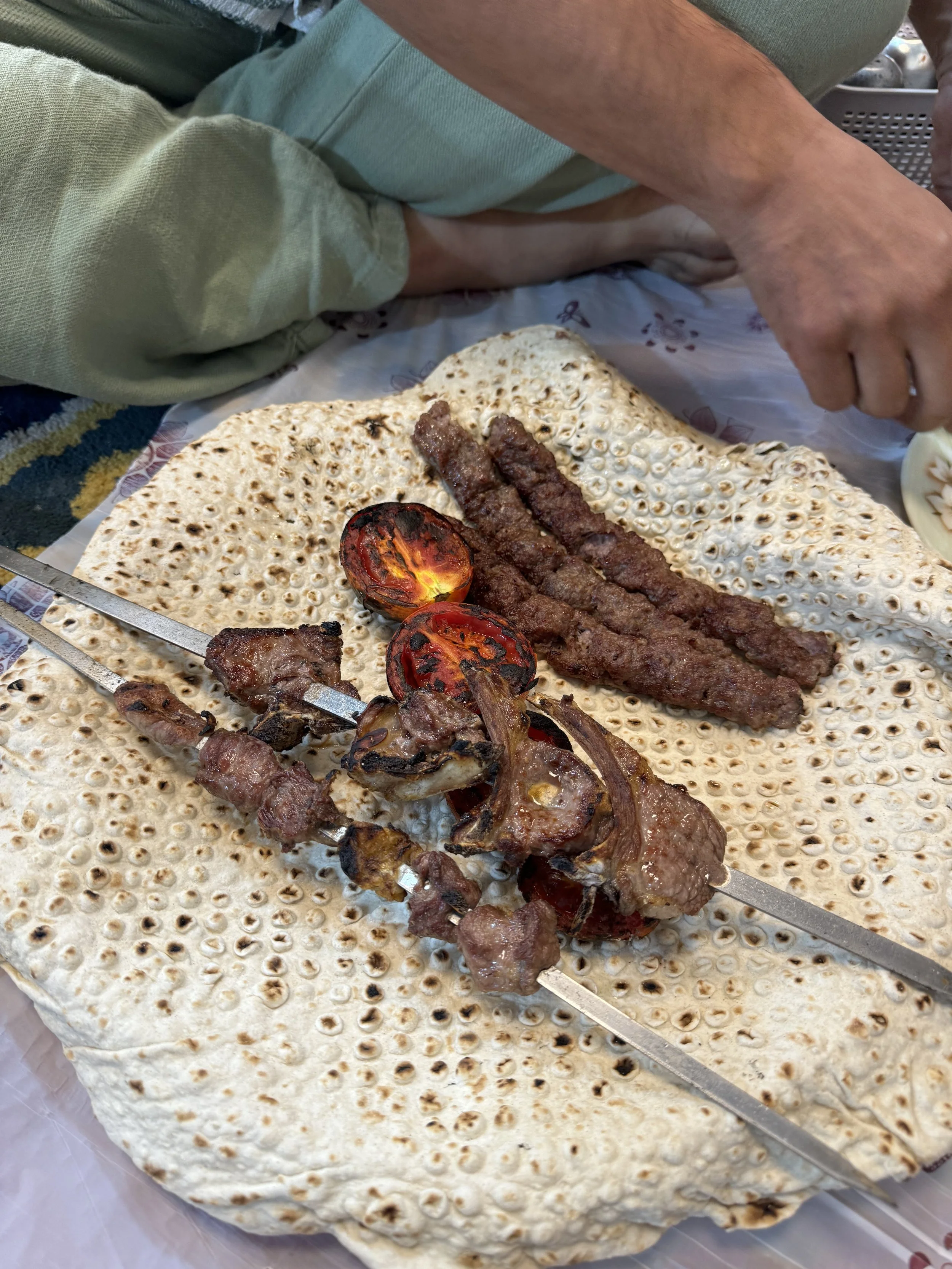 Close-up of skewered grilled meat on a traditional flatbread, with grilled tomatoes and additional meat strips, on a person's lap.