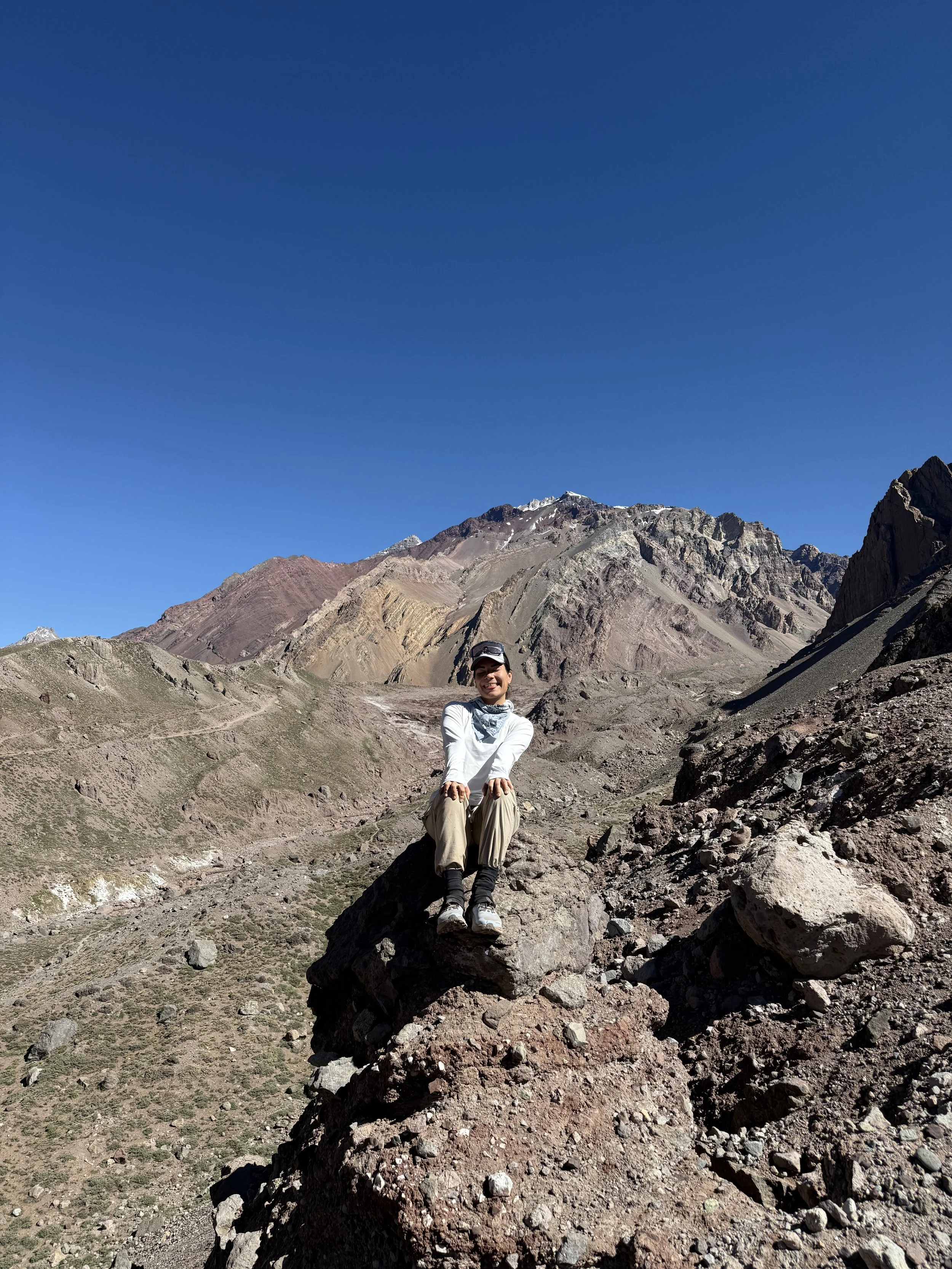 A person sitting on a large rock in a mountainous desert landscape with colorful layered mountains and a clear blue sky.