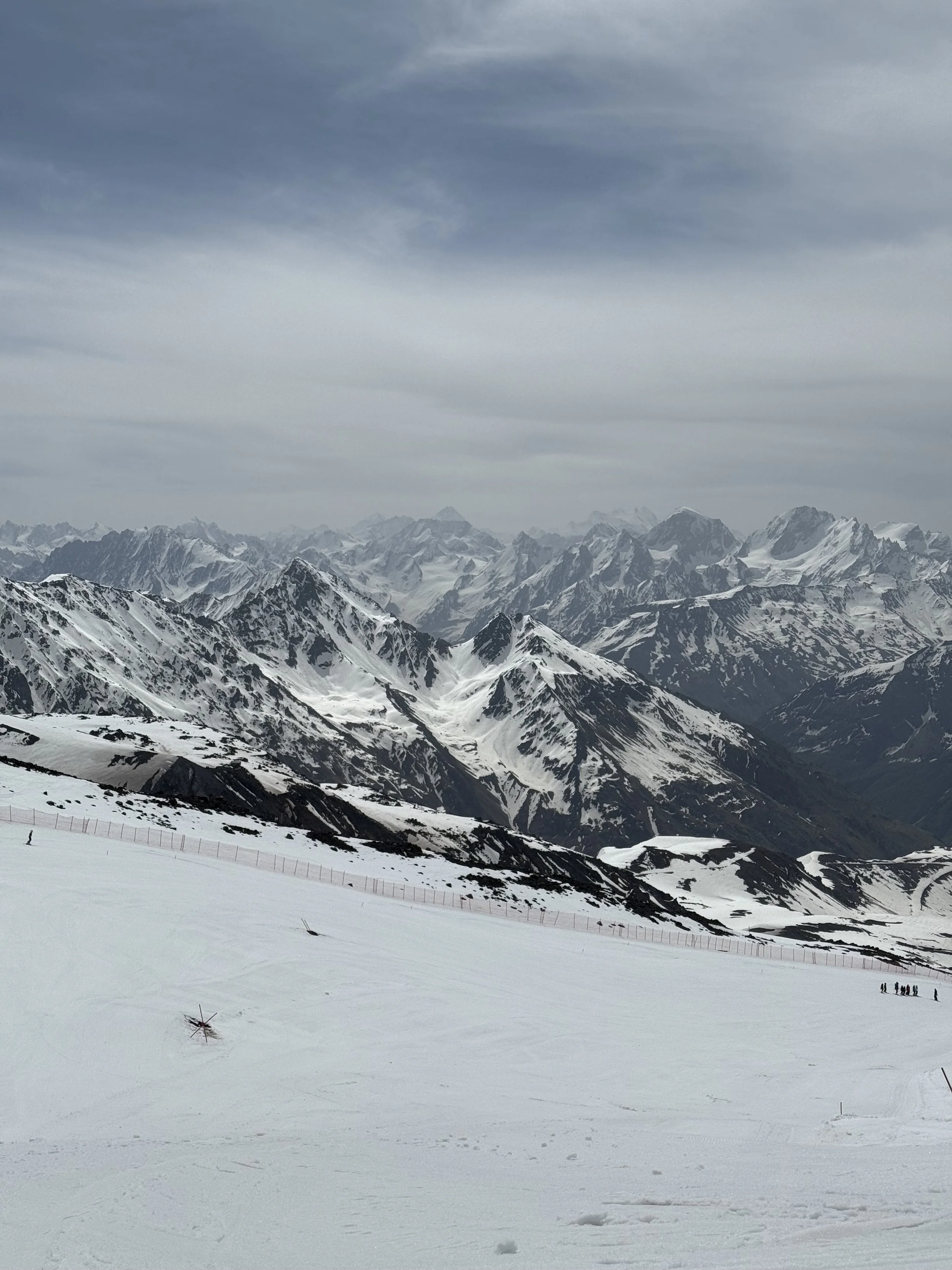 Snow-covered mountains under a cloudy sky with a flat snowy foreground and a few distant figures.