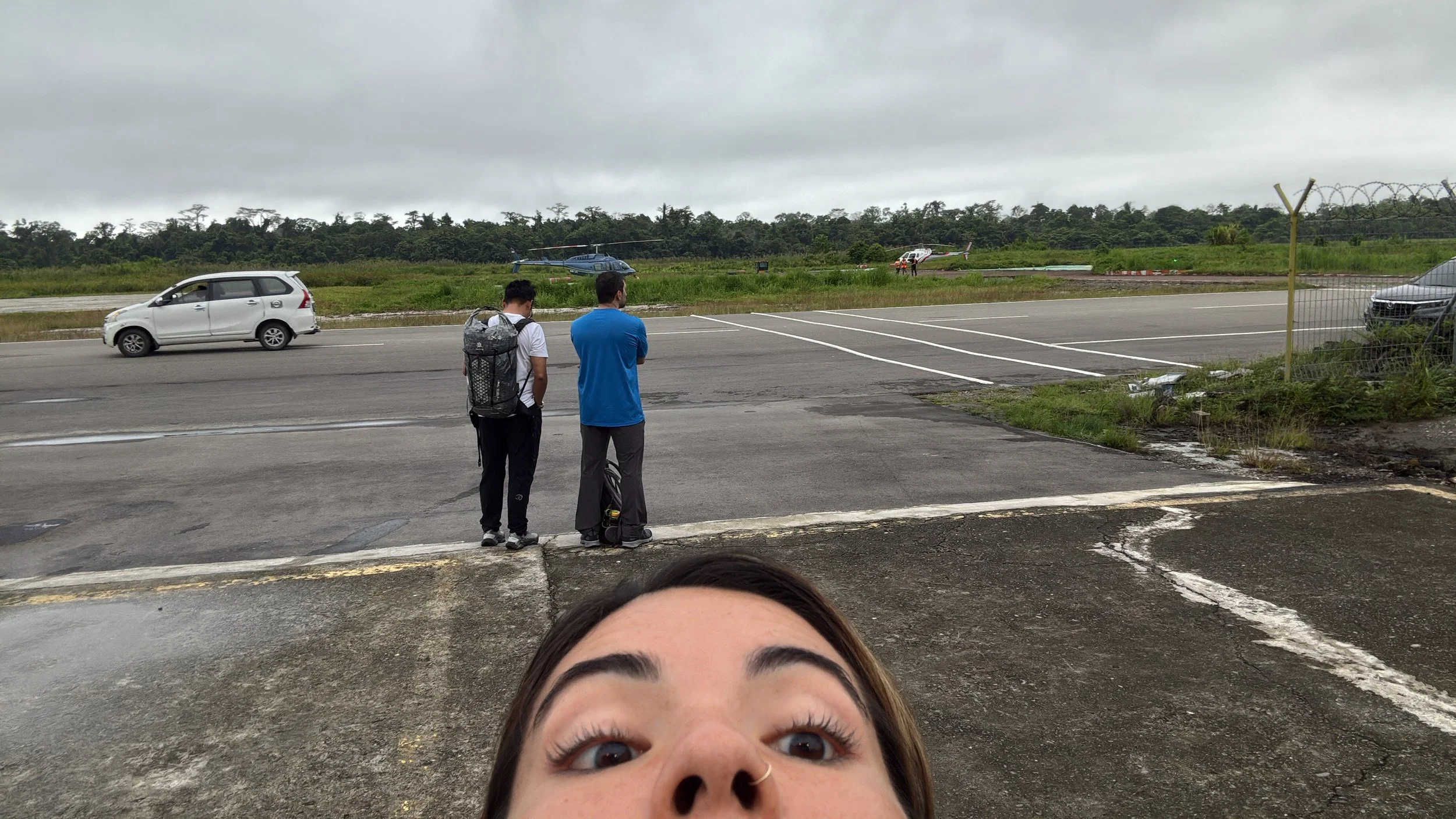 People standing on the side of a road, watching helicopters land and take off on a grassy airstrip on a cloudy day.