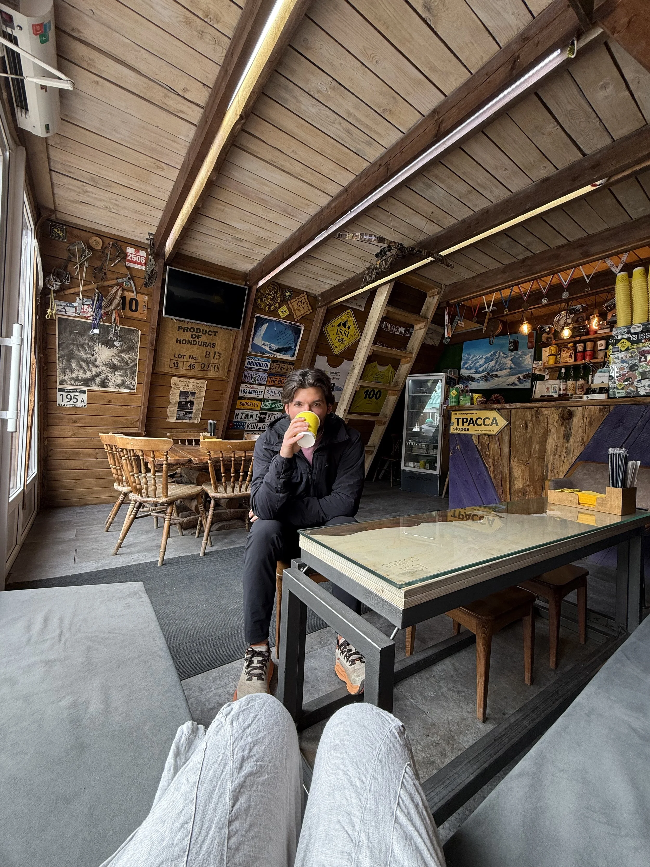 A person sitting and drinking a yellow mug inside a cozy, rustic wooden cafe or lodge, with wood-paneled walls decorated with signs, artwork, and memorabilia. The photo is taken from a first-person perspective, showing the photographer's legs in the
