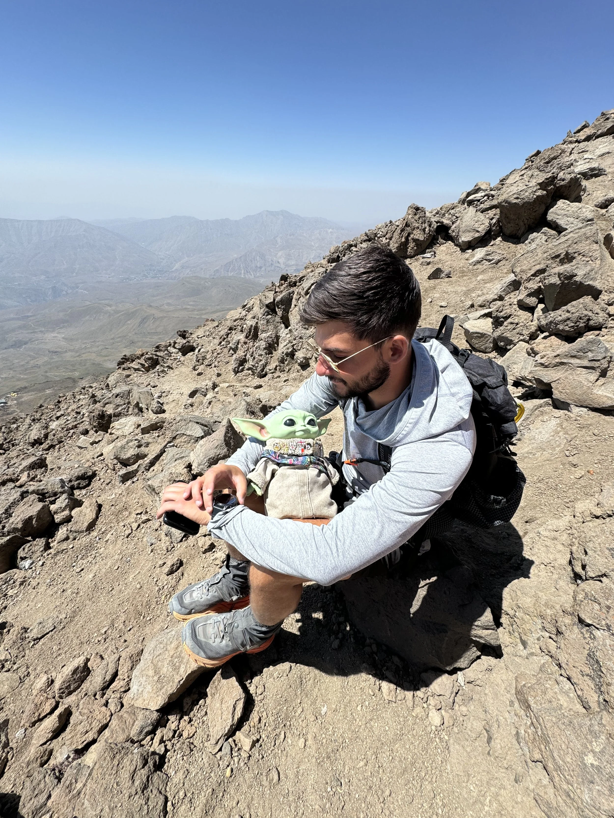 A man sitting on a rock on a mountain trail, holding a small plush toy of Baby Yoda, with a rugged mountainous landscape and a clear blue sky in the background.