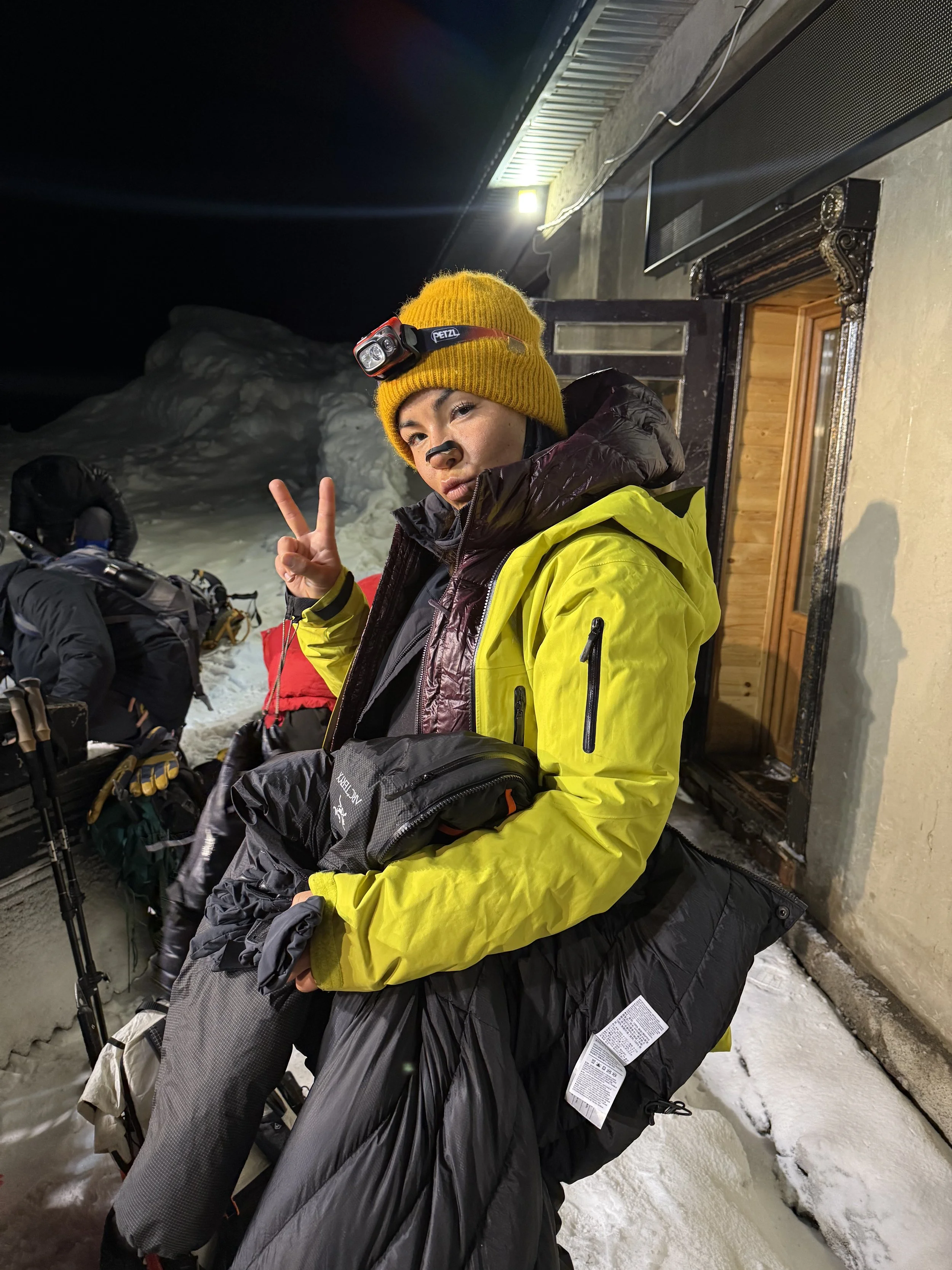 Person dressed in winter gear, wearing a yellow jacket and a yellow knit hat with a headlamp, making a peace sign at night with snow and a mountain in the background.