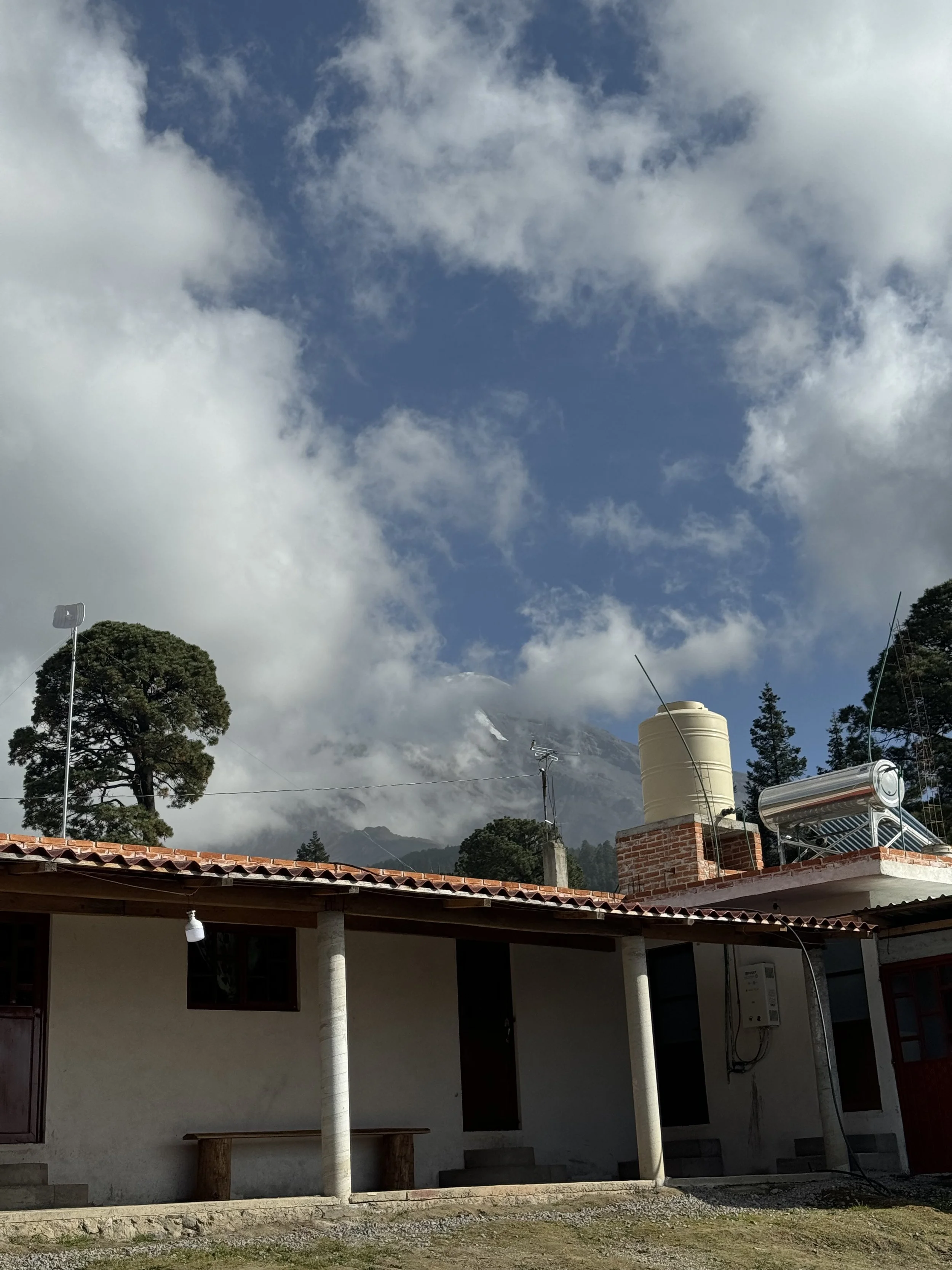 Mountain with clouds, trees, and rooftops of a house with water tanks.