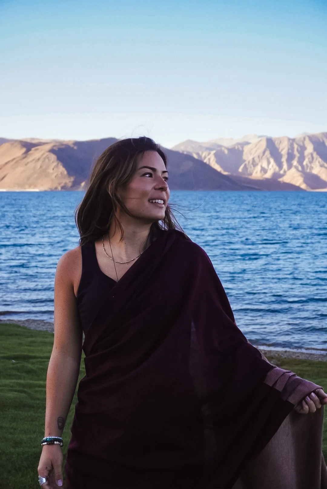 A woman standing near a lake with mountains in the background, smiling and holding her sari.