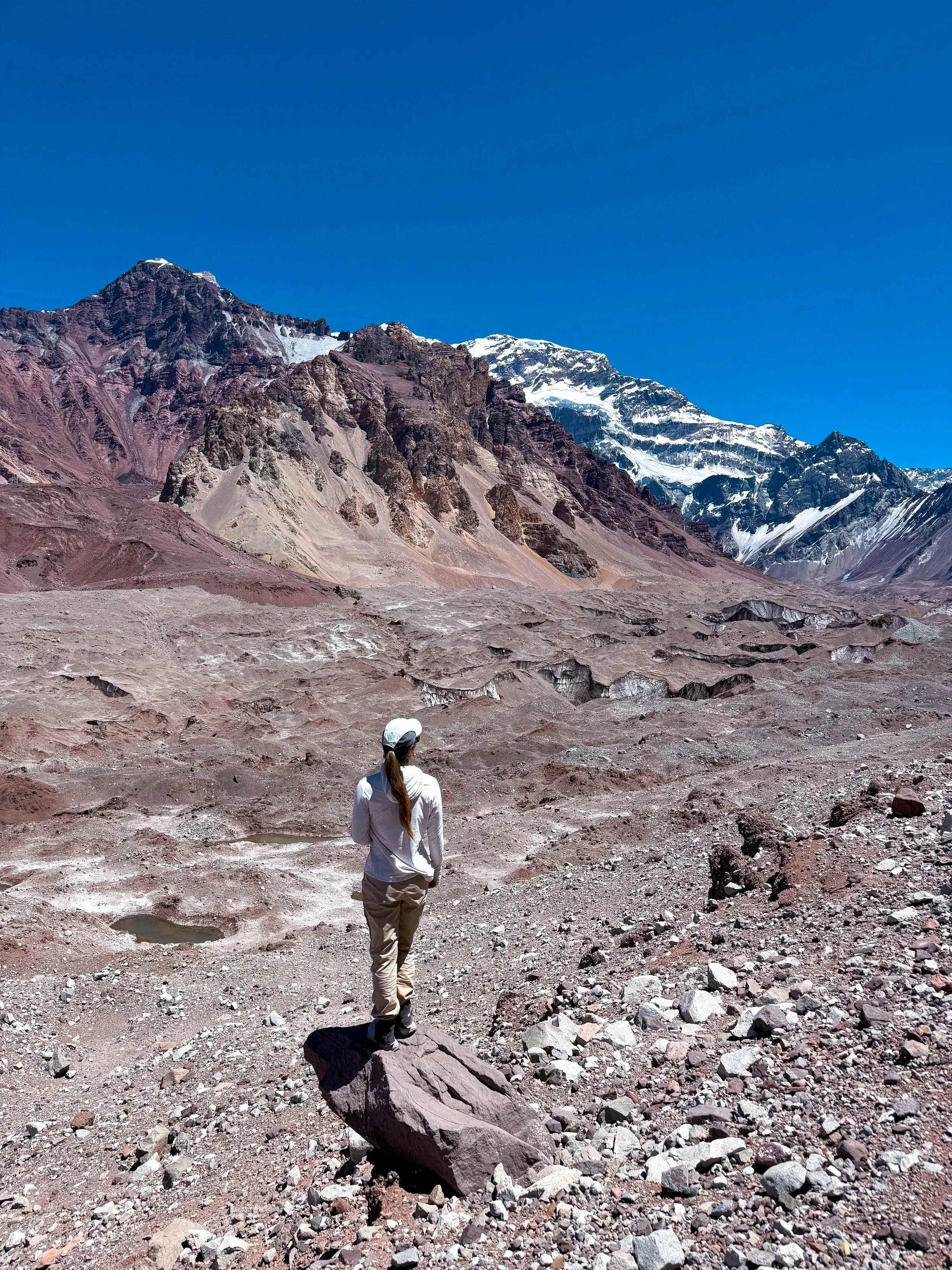 A woman stands on a large rock in a barren, rocky landscape with mountains and snow-covered peaks in the background under a clear blue sky.