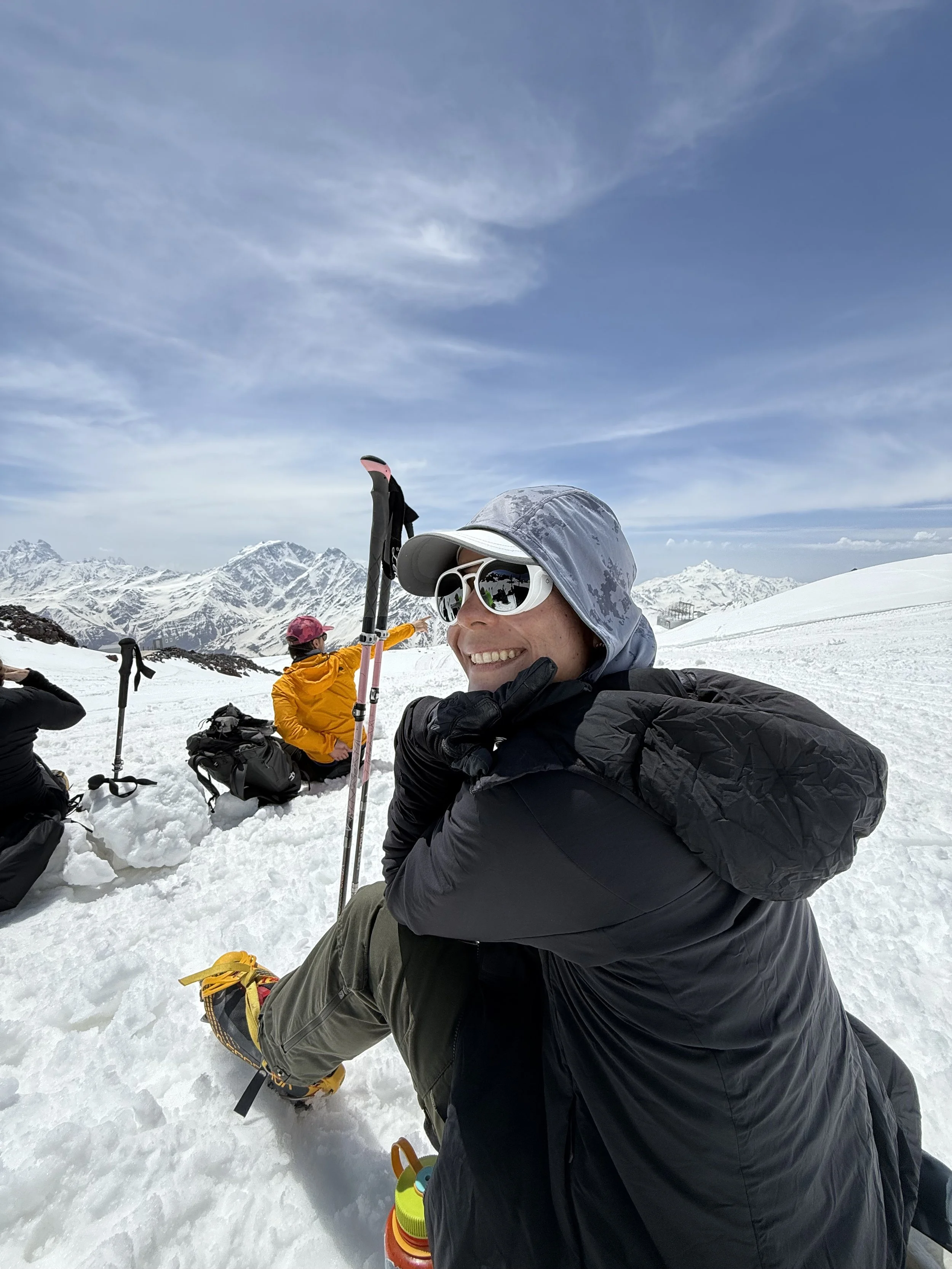 A woman smiling and sitting in the snow in a snowy mountain landscape, wearing sunglasses, a hooded jacket, and a hat, with other people and mountain peaks in the background.