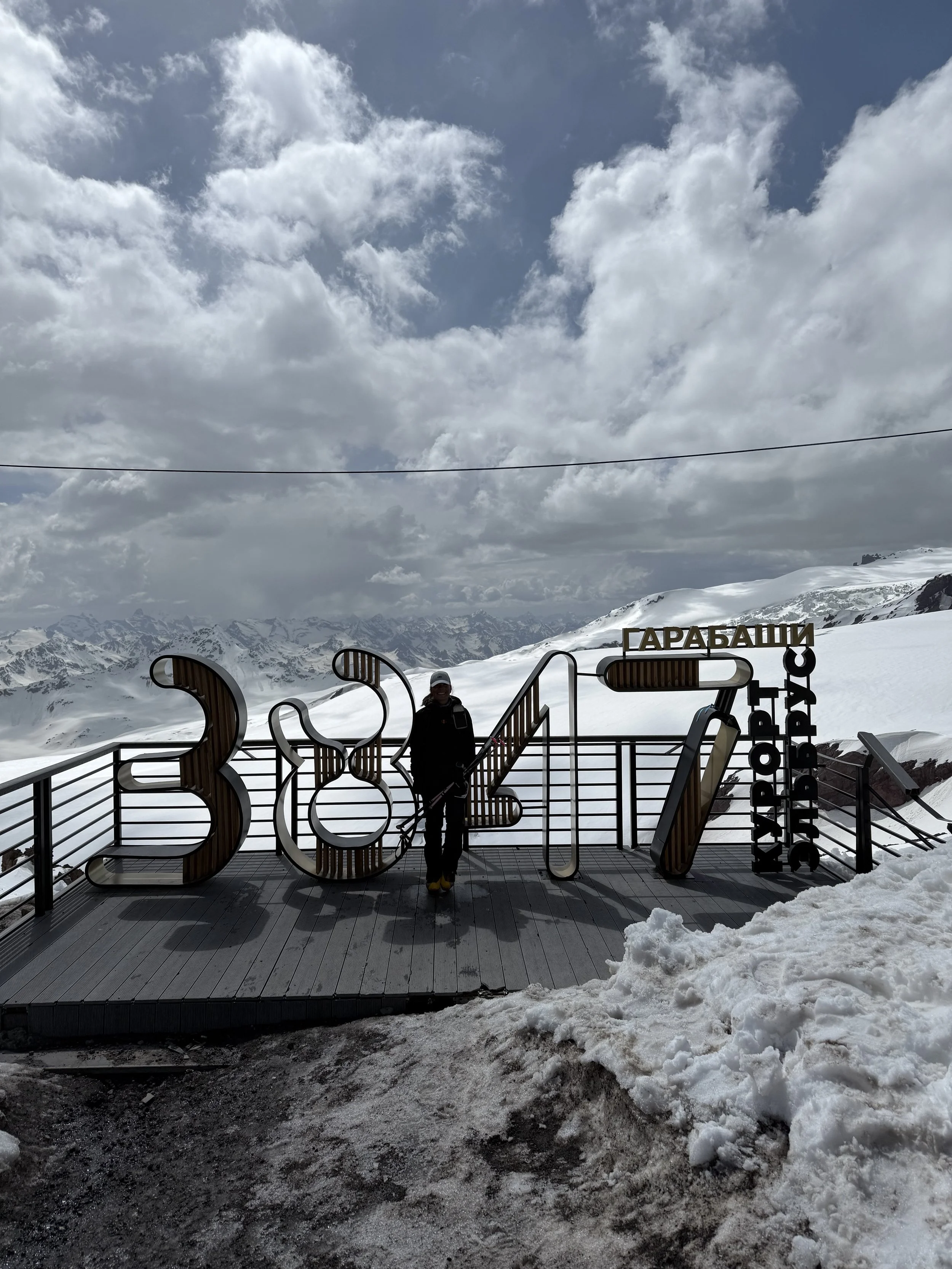 A person standing on a viewing platform with large decorative numbers and signs, overlooking snow-covered mountains under a cloudy sky.