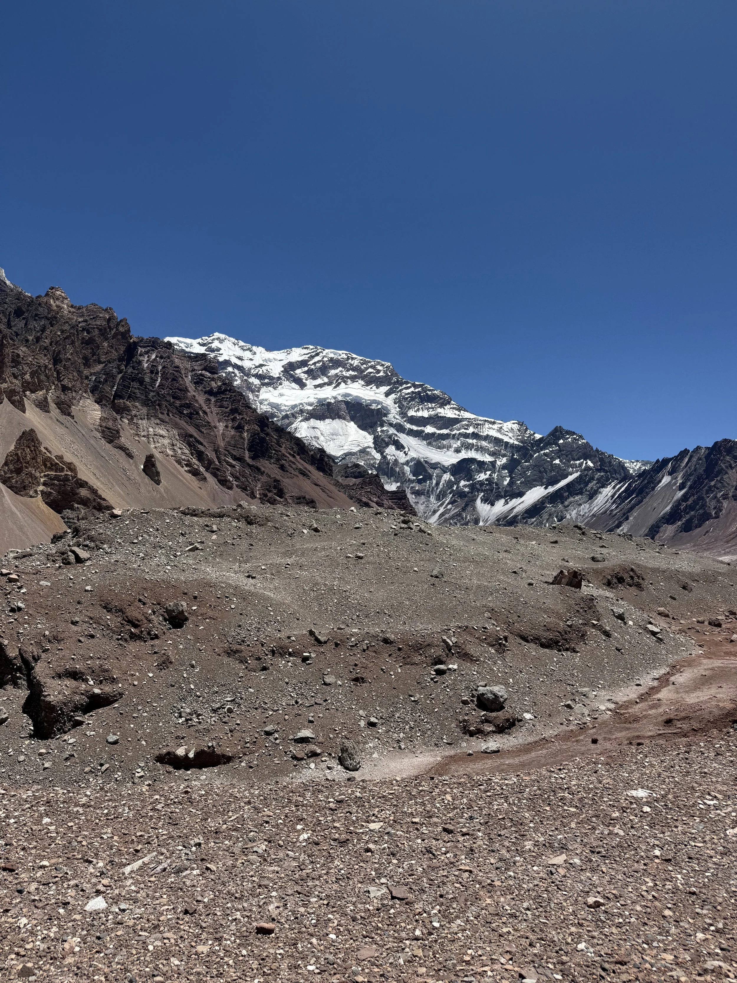 A rugged dirt trail winds through a barren, rocky terrain with mountains in the background, some covered in snow, under a clear blue sky.