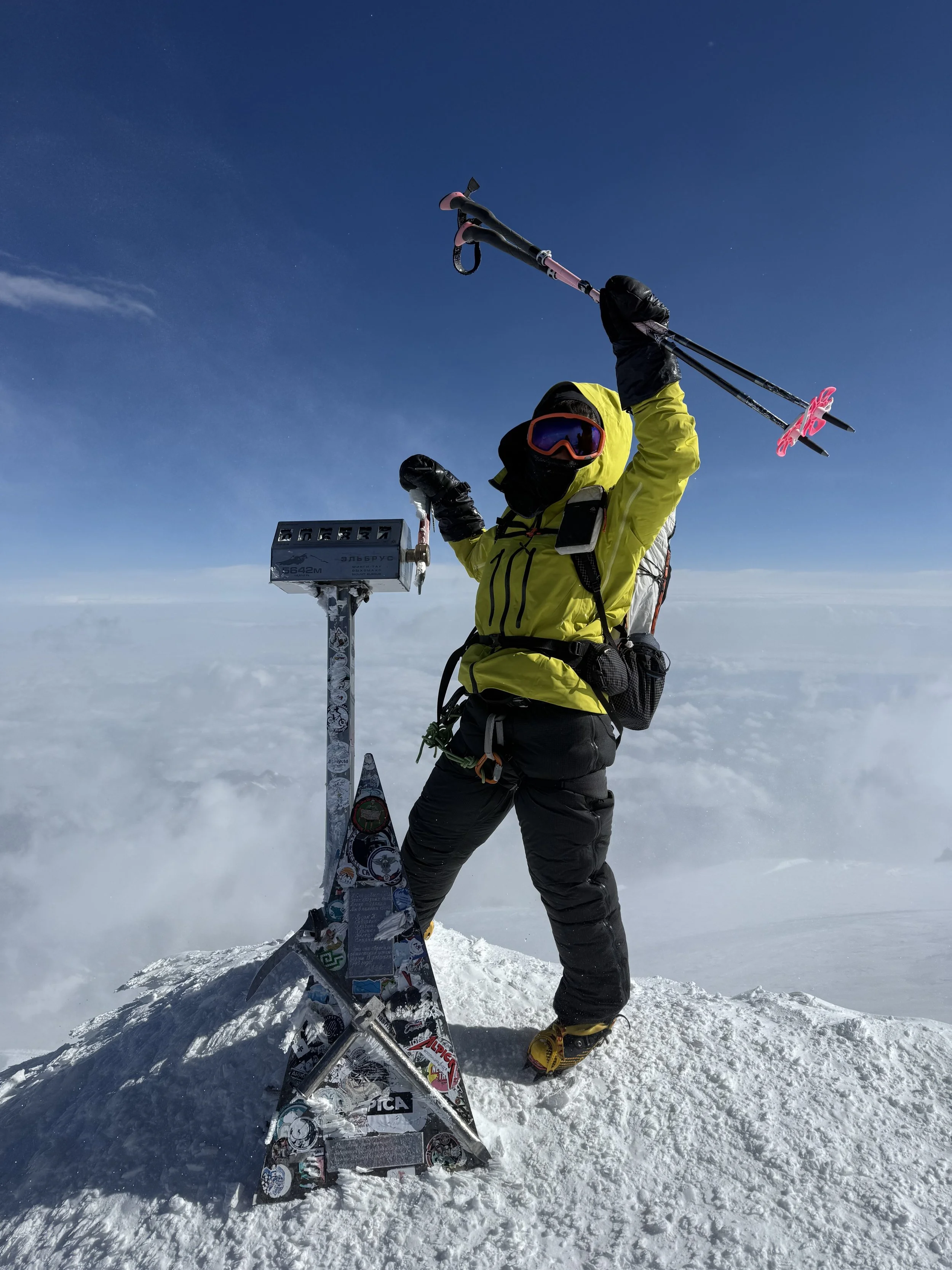 Climber standing on snowy mountain peak holding trekking poles above their head with a colorful sticker-covered snowboarding snowboard leaning against a weather station on the peak, against a backdrop of clouds and clear blue sky.