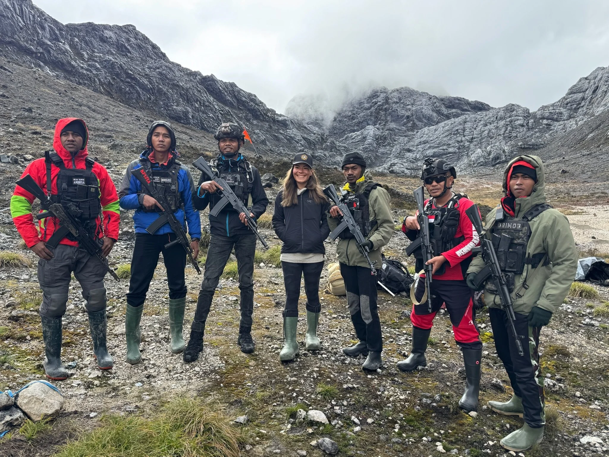 Group of six people dressed in outdoor gear and tactical vests, holding rifles, standing on rocky terrain with mountains in the background.