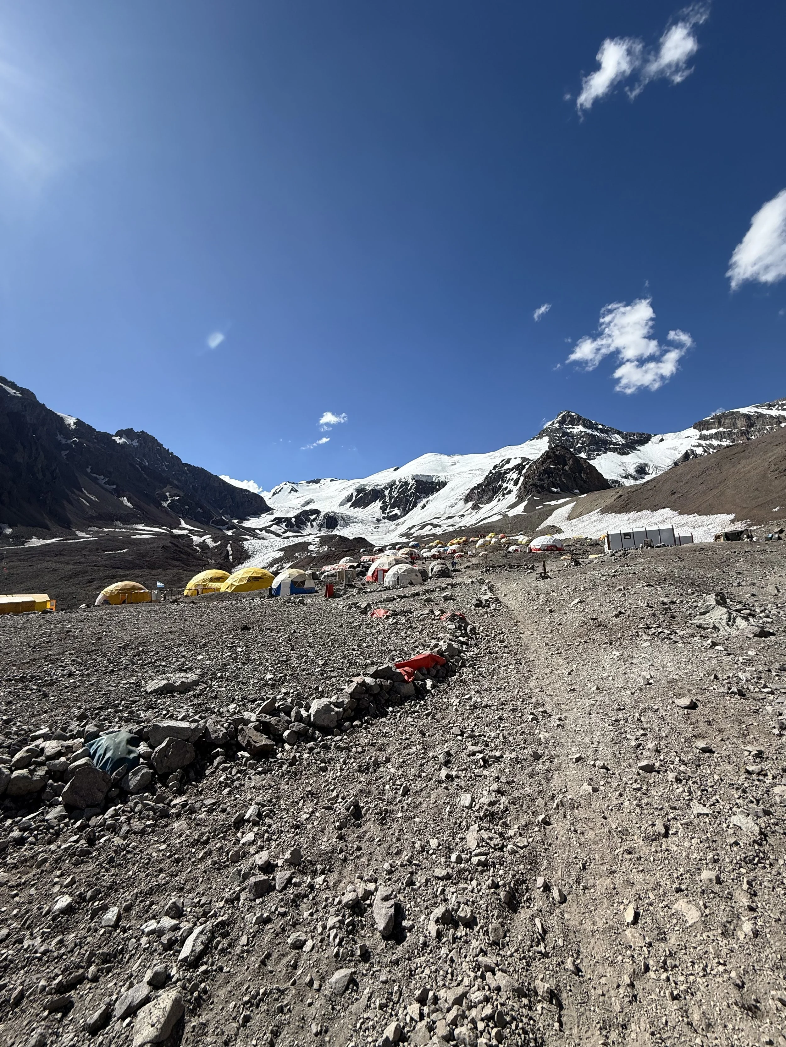 Mountains with snow, rocky dirt trail, and a campsite with tents in a high-altitude area under a blue sky.