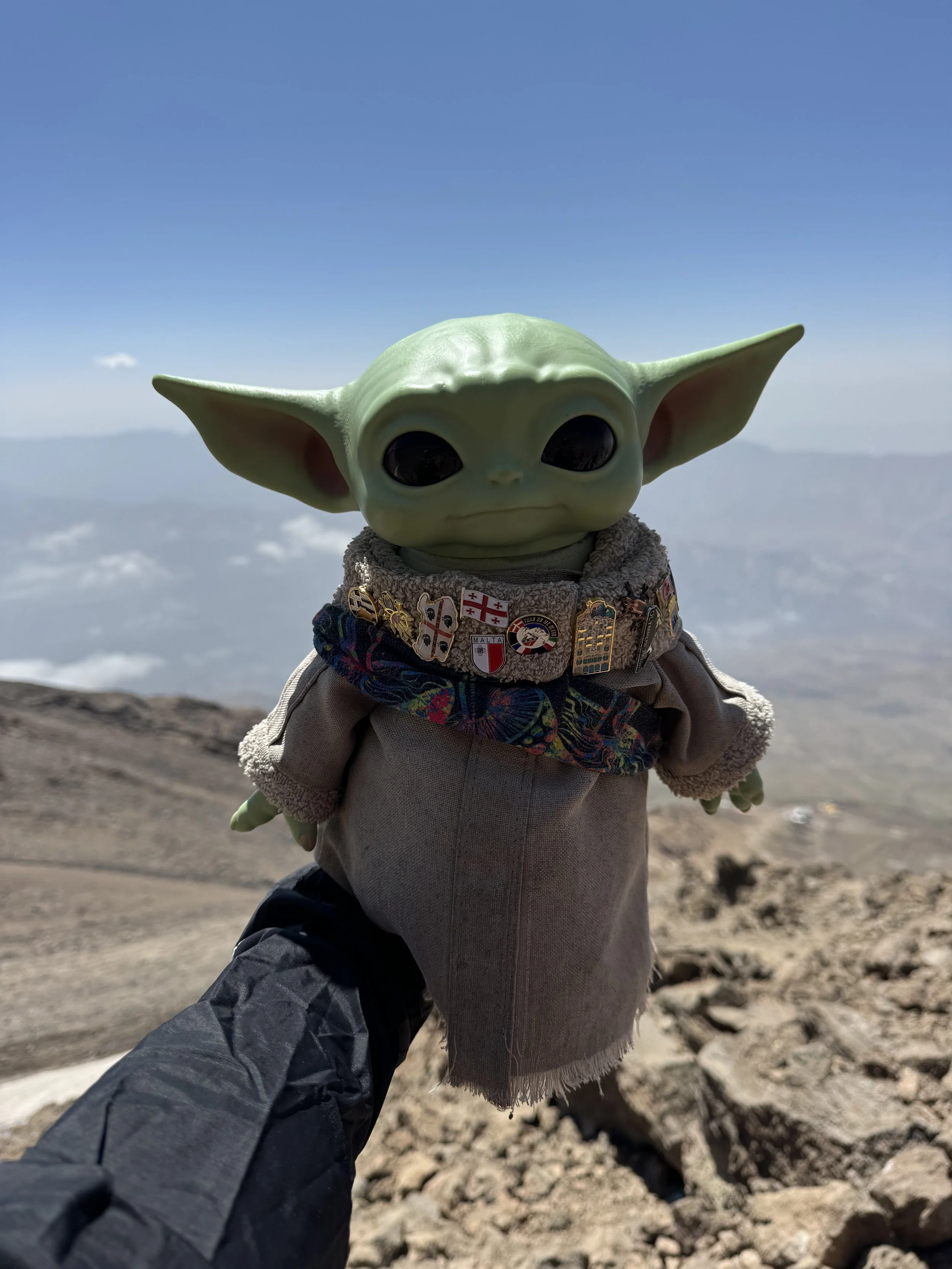 A toy figure of Baby Yoda (Grogu) from Star Wars, dressed in a beige outfit with a decorated collar, being held up against a cloudy sky and mountainous landscape.