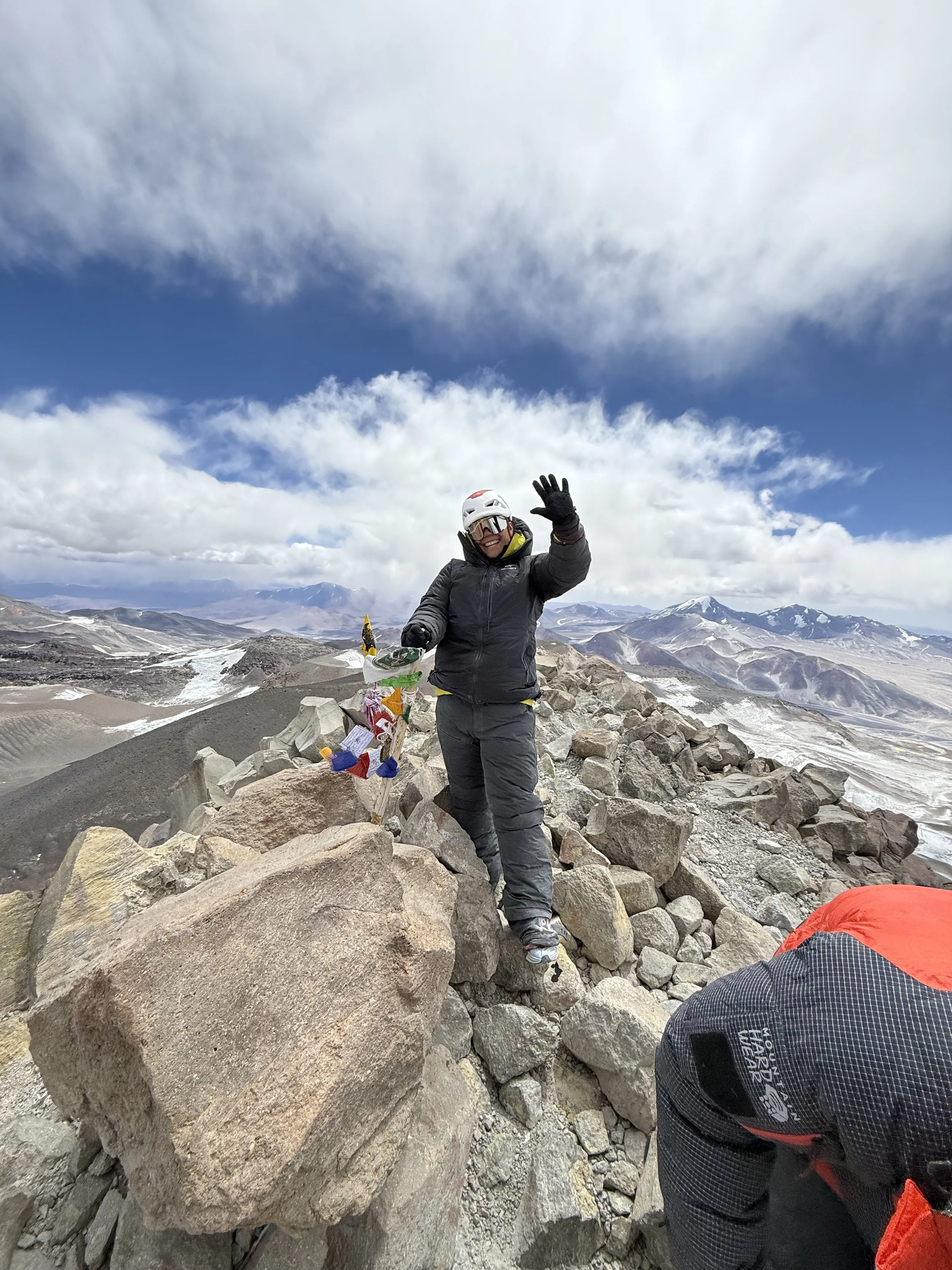 Person in mountaineering gear hiking on rocky mountain summit, smiling and waving, with scenic mountain landscape and partly cloudy sky in the background.