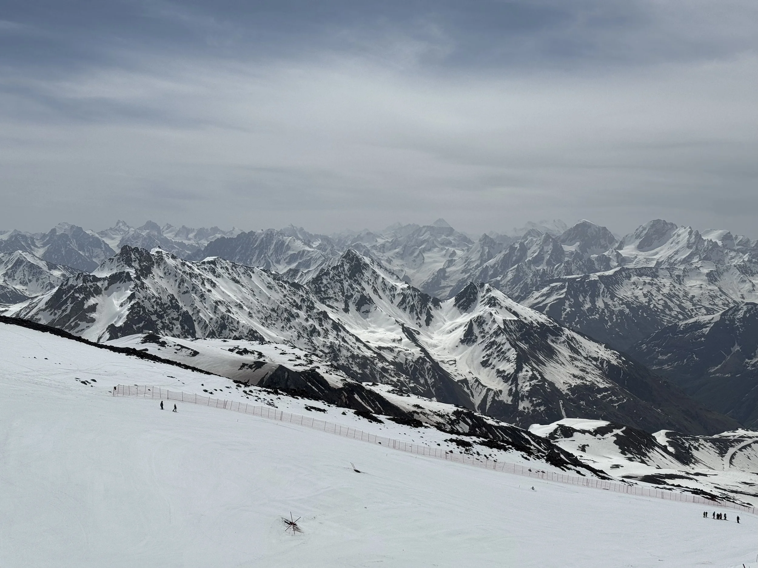 Snow-covered mountain range with rugged peaks under cloudy sky, with a few people visible on a snowy slope in the foreground.