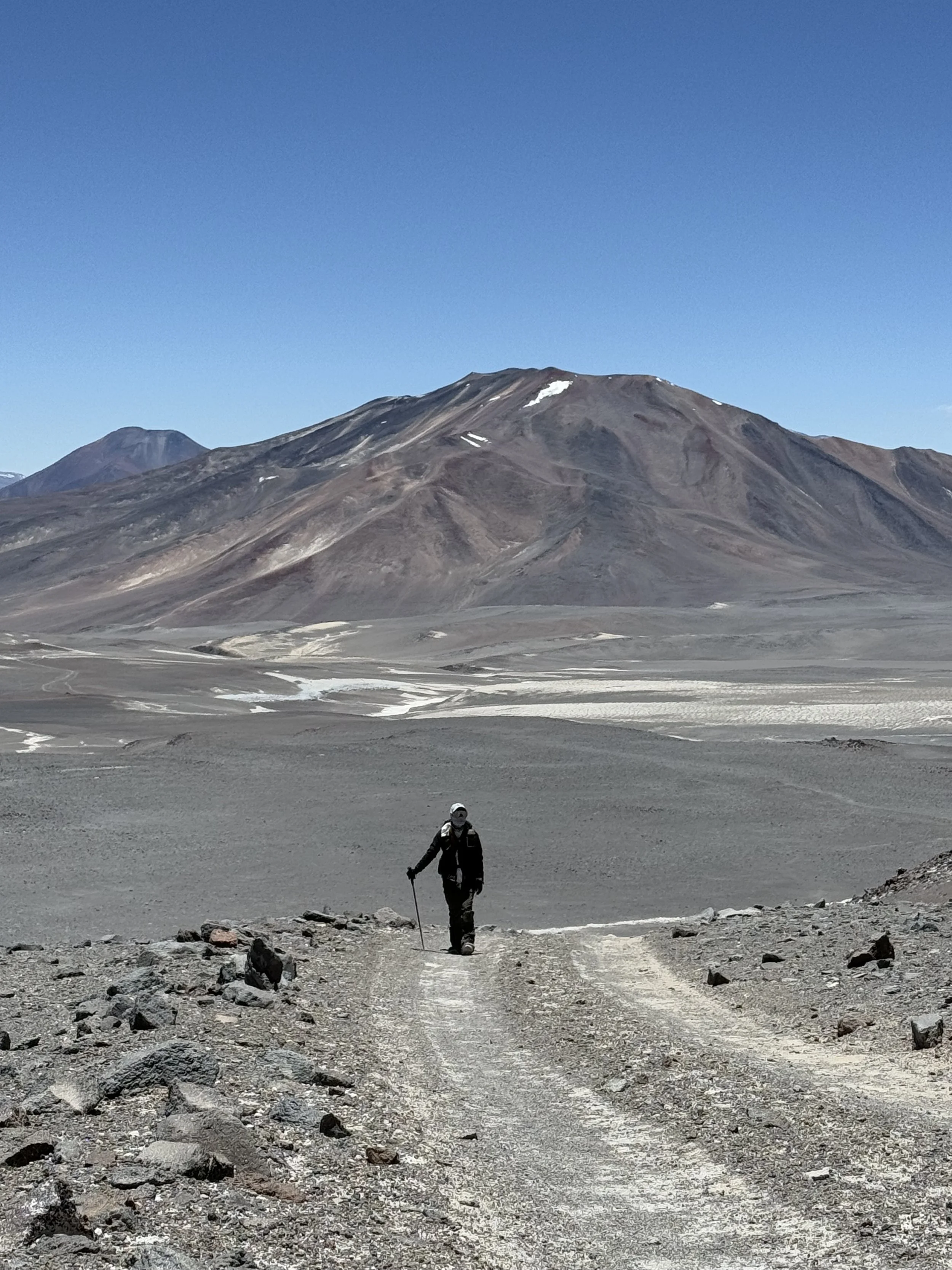 A person hiking on a dirt trail in a barren, mountainous landscape with a mountain in the background, under a clear blue sky.