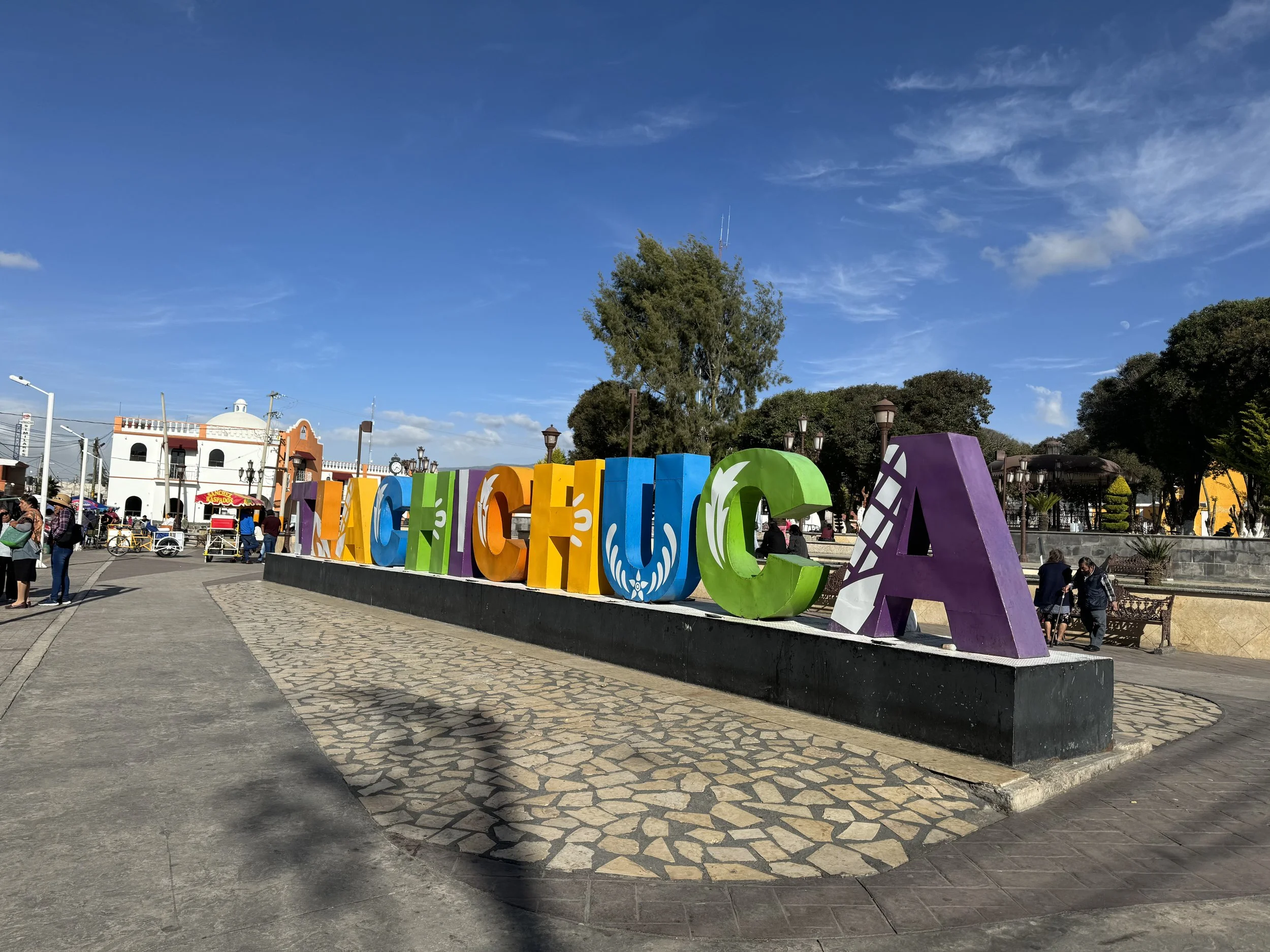Colorful large letters spelling 'VALLADO CUA' on a black platform in a public square with a blue sky, trees, and people in the background.