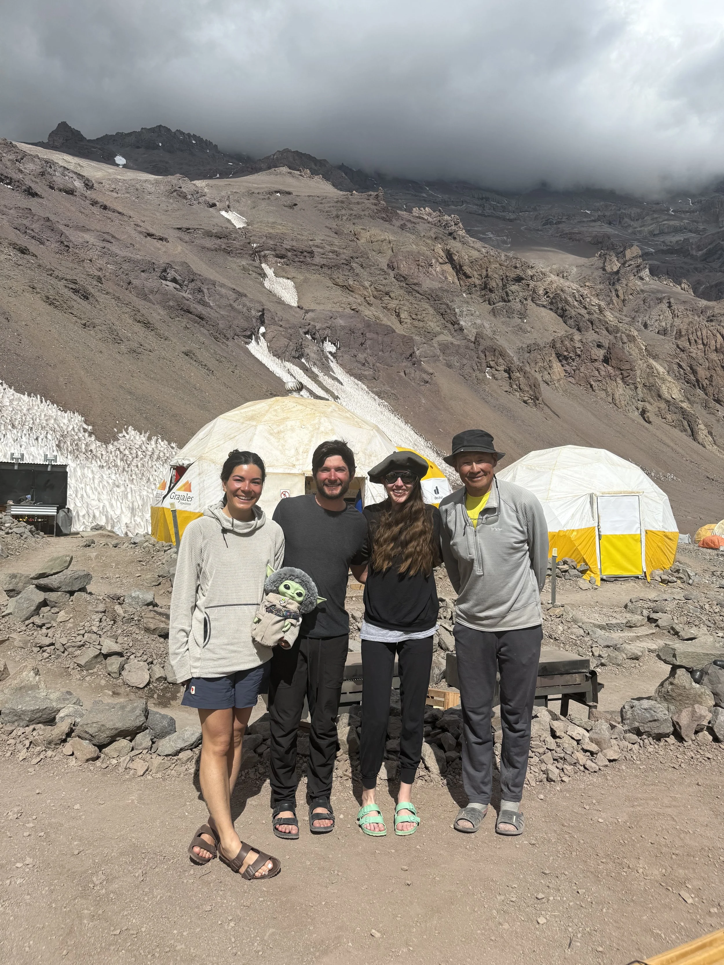 Four people standing together in front of a mountainous volcanic landscape with tents, rocky terrain, and snow patches.