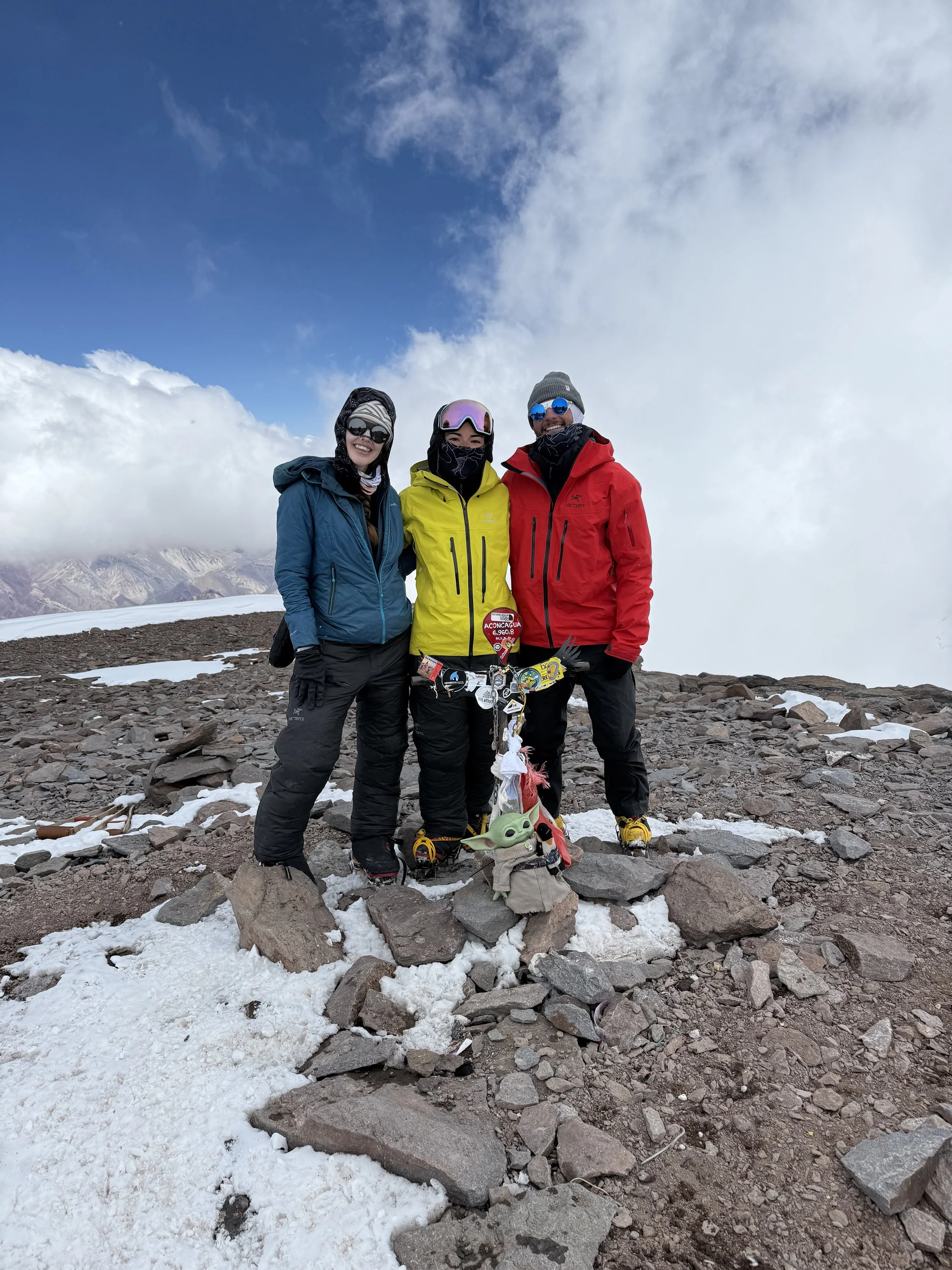 Three people in winter hiking gear standing on rocky, snowy mountain terrain with cloudy sky and mountain view in the background.