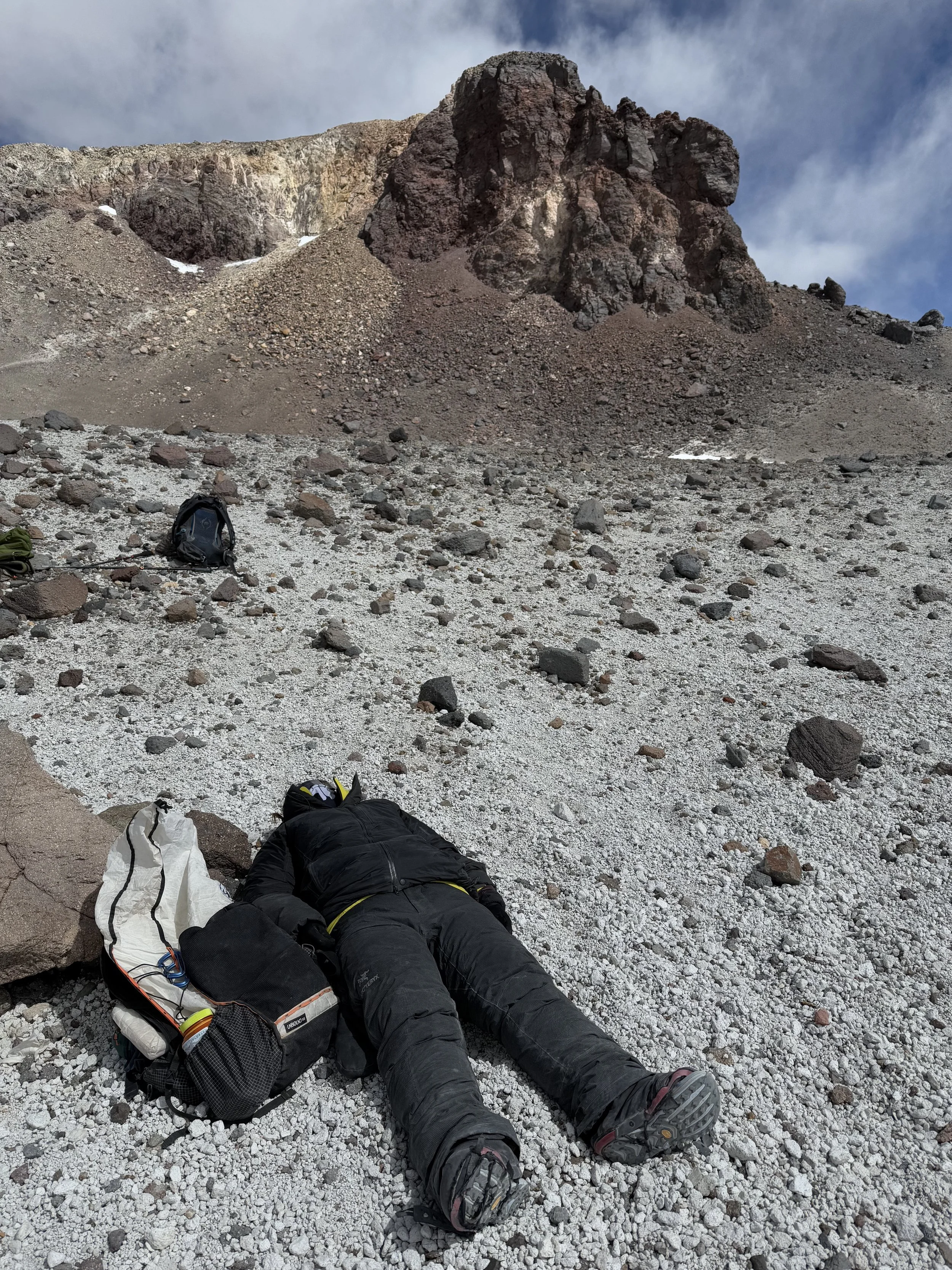 A person lying on the rocky ground of a mountain slope, wearing black outdoor gear and surrounded by backpacks, with a large rocky mountain formation in the background under a partly cloudy sky.