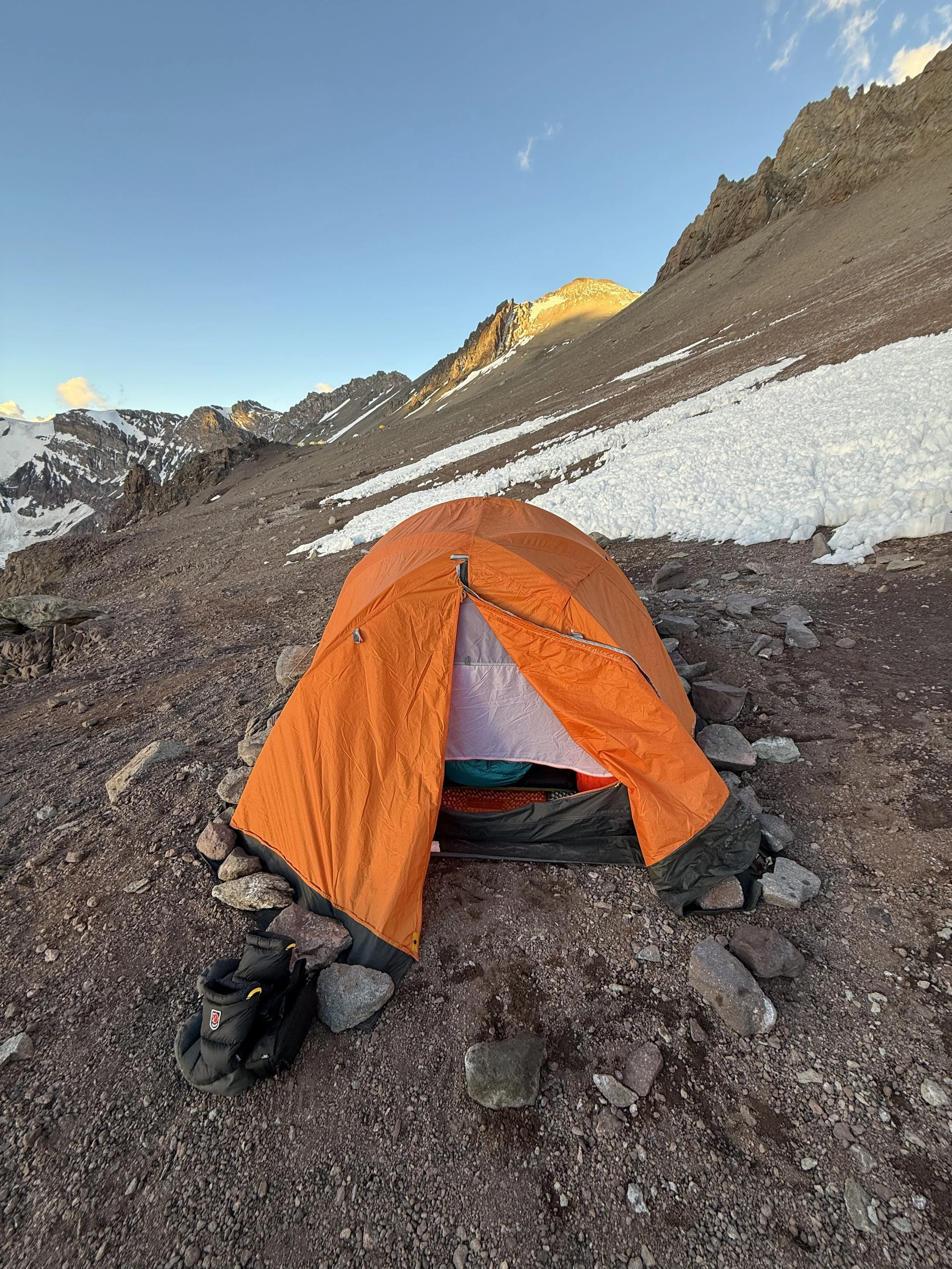 Orange camping tent on rocky ground in a mountainous area with patches of snow, under a clear blue sky.