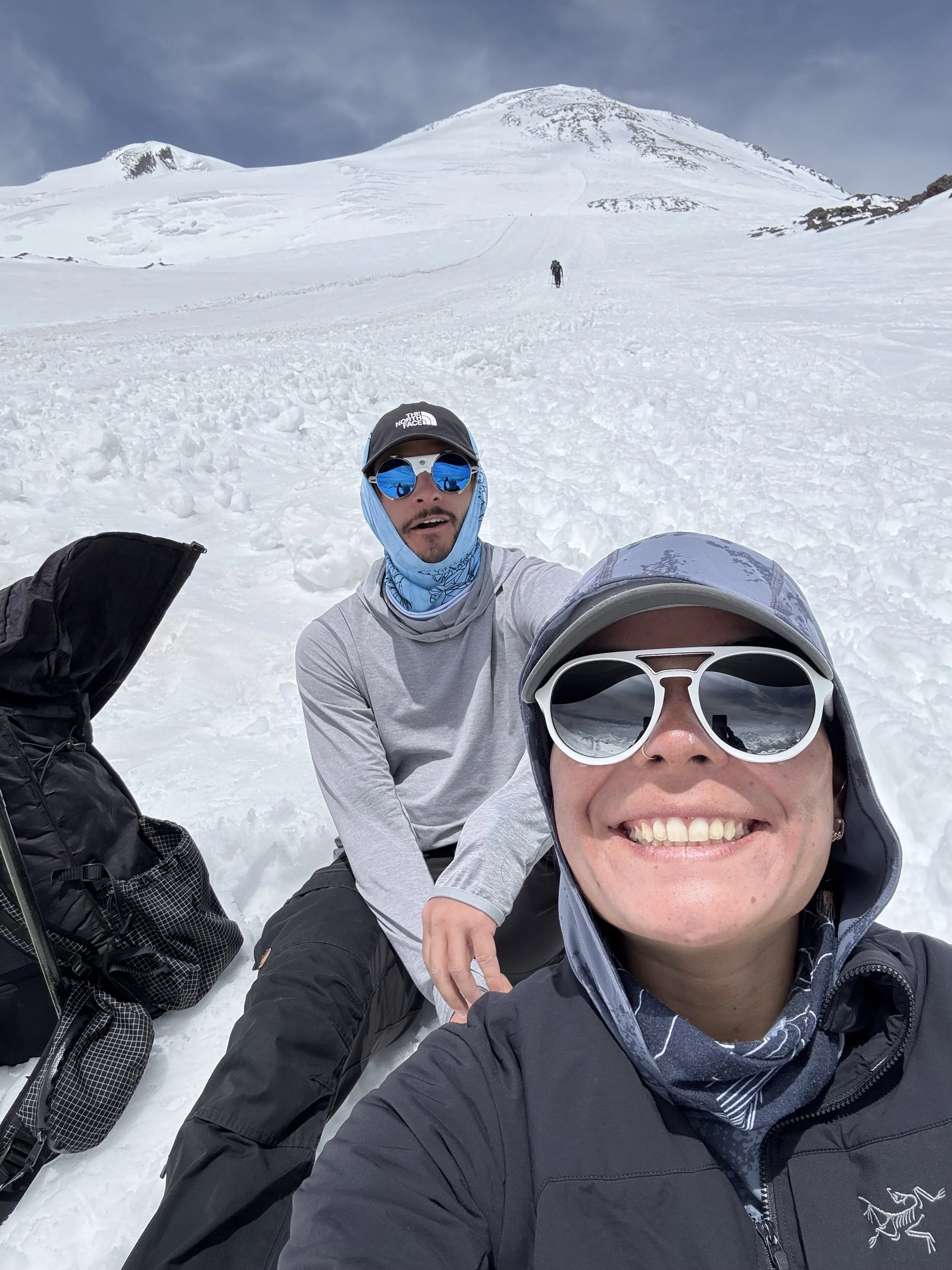 Two people wearing sunglasses and outdoor gear sitting in the snow, with a snowy mountain in the background.