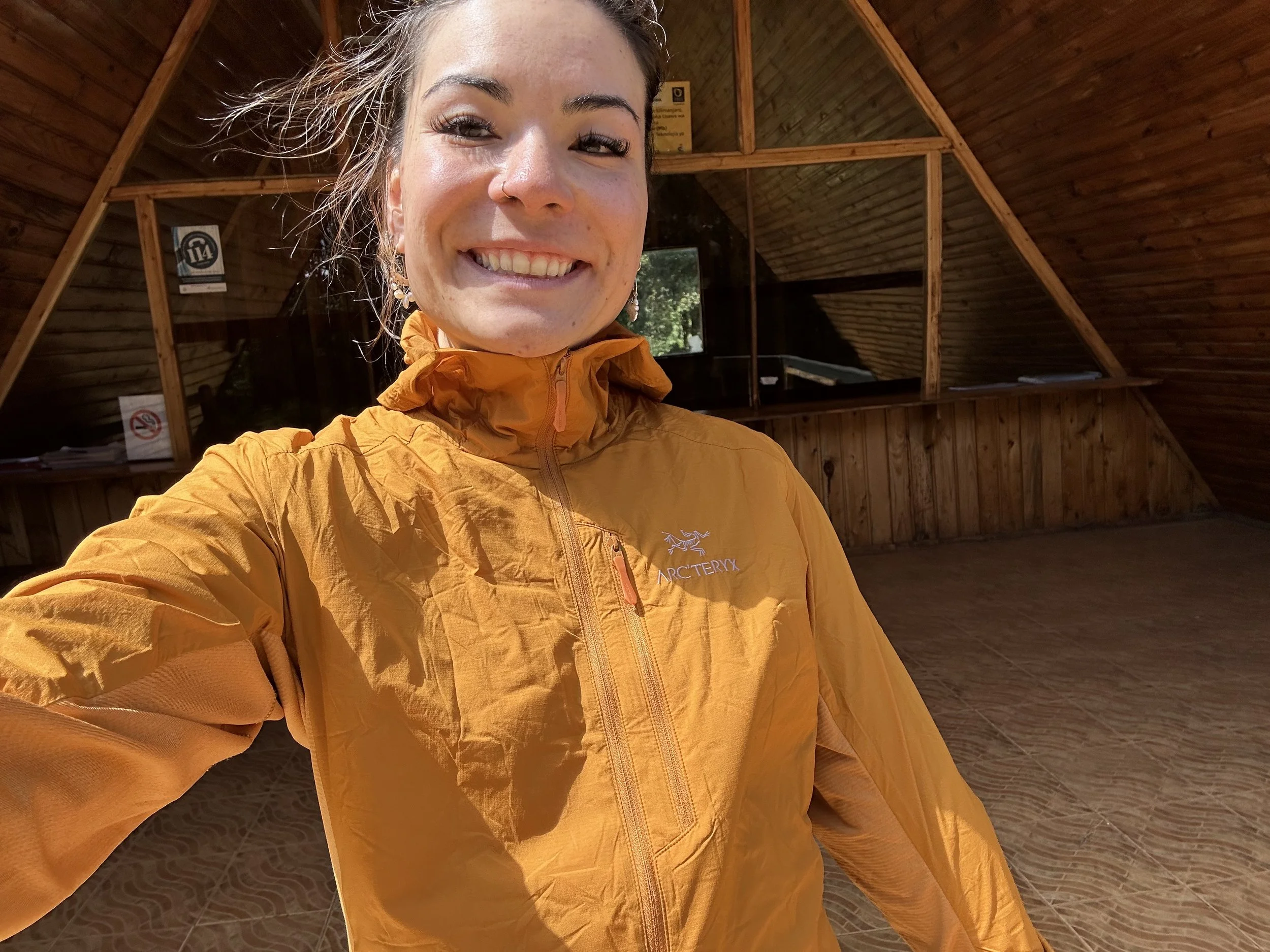 A woman takes a selfie inside a wooden, attic-like room with sloped walls. She is smiling, has dark hair, and is wearing an orange Arc'teryx jacket.