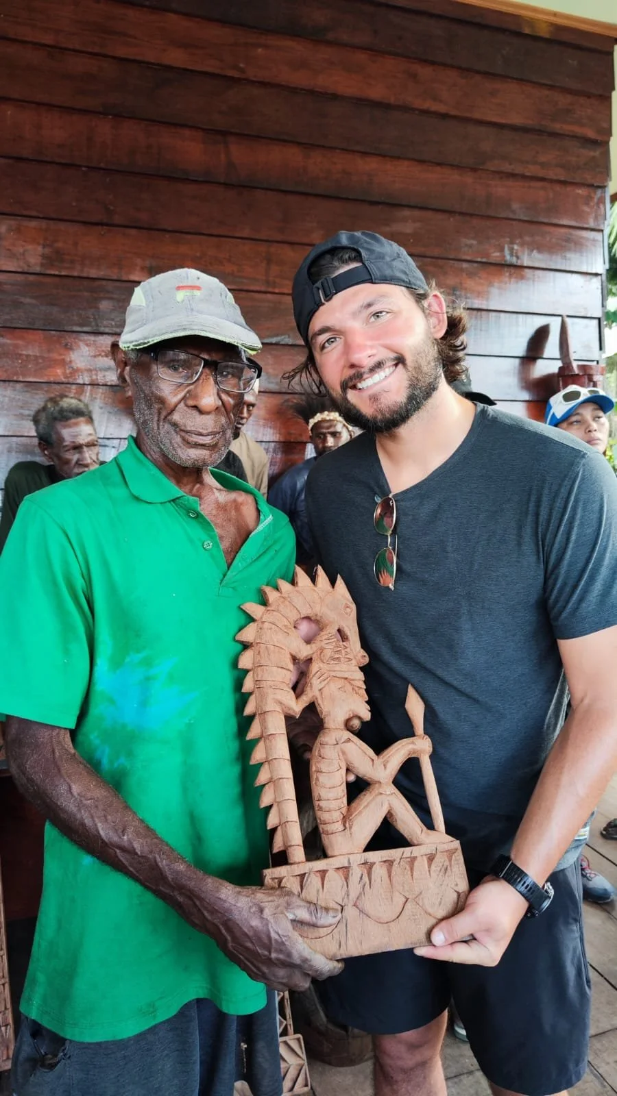 An elderly man with a green shirt, glasses, and a cap holding a carved wooden sculpture of a seated figure with a headdress, next to a smiling young man in a black t-shirt, black cap, and sunglasses hanging from his shirt, standing in front of a wood