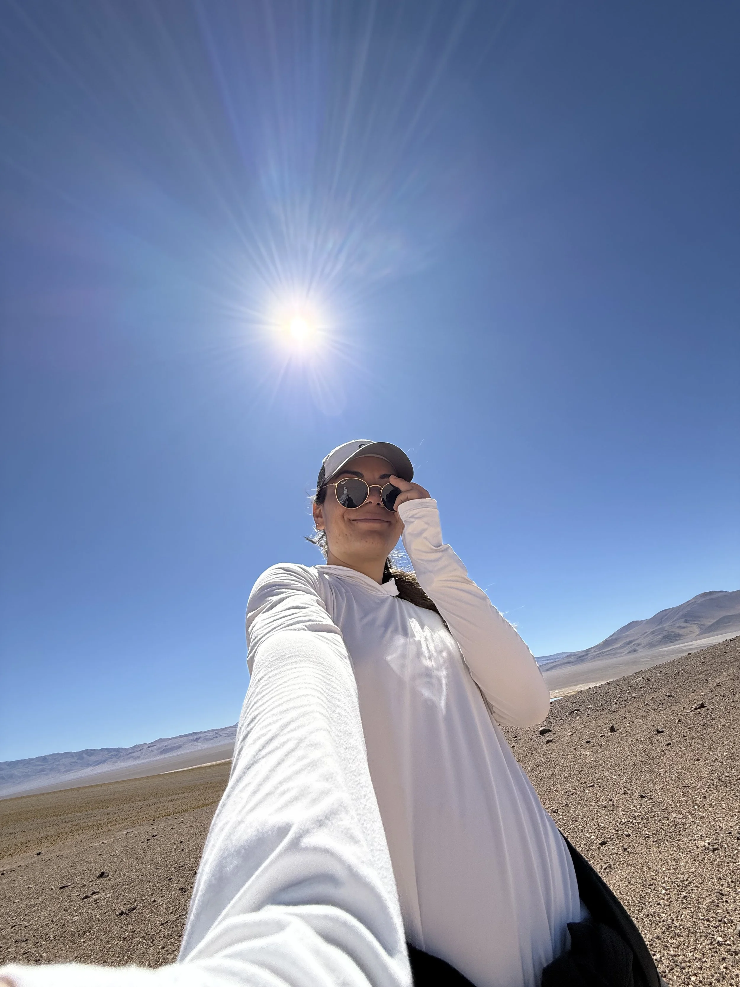 A person in a white long-sleeve shirt, sunglasses, and a cap taking a selfie in a desert landscape with mountains in the background under a bright sun in clear blue sky.