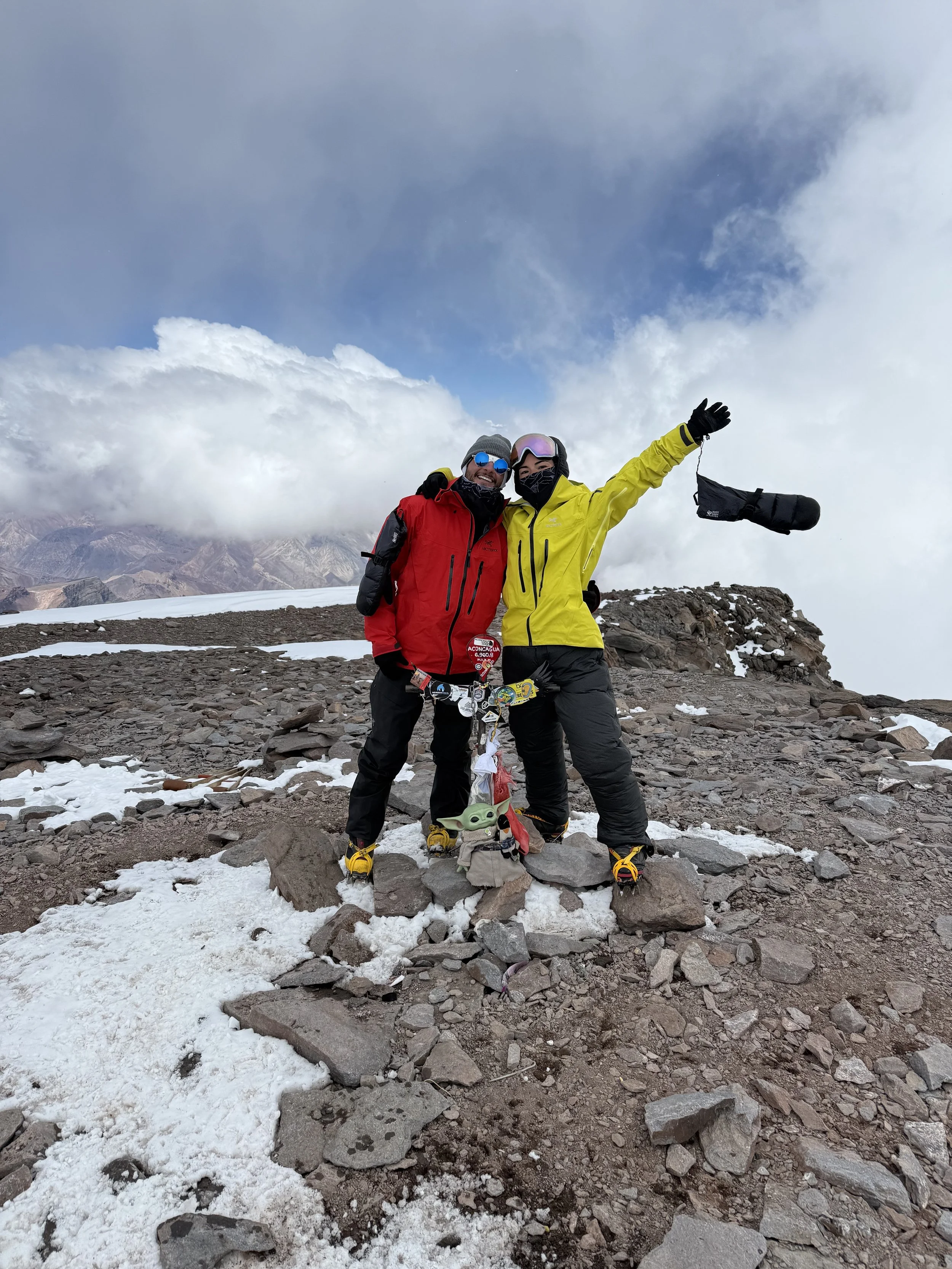 Two climbers in colorful gear standing on rocky mountain summit with snow patches, celebrating with arms raised, against cloudy sky and distant mountain range.