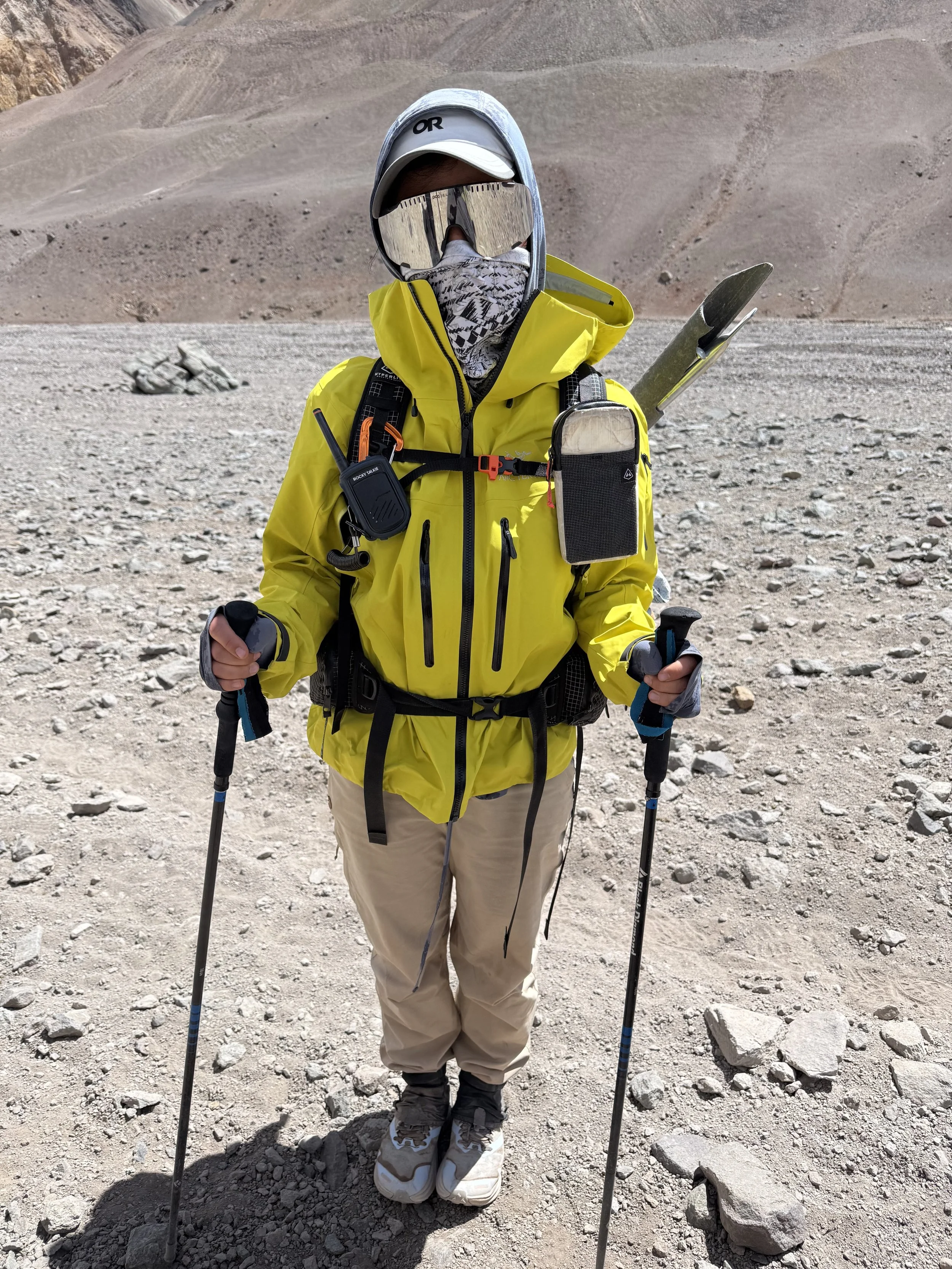 Person hiking in a mountainous, rocky landscape, wearing a bright yellow jacket, beige pants, a cap, goggles, and carrying trekking poles and a backpack with equipment.
